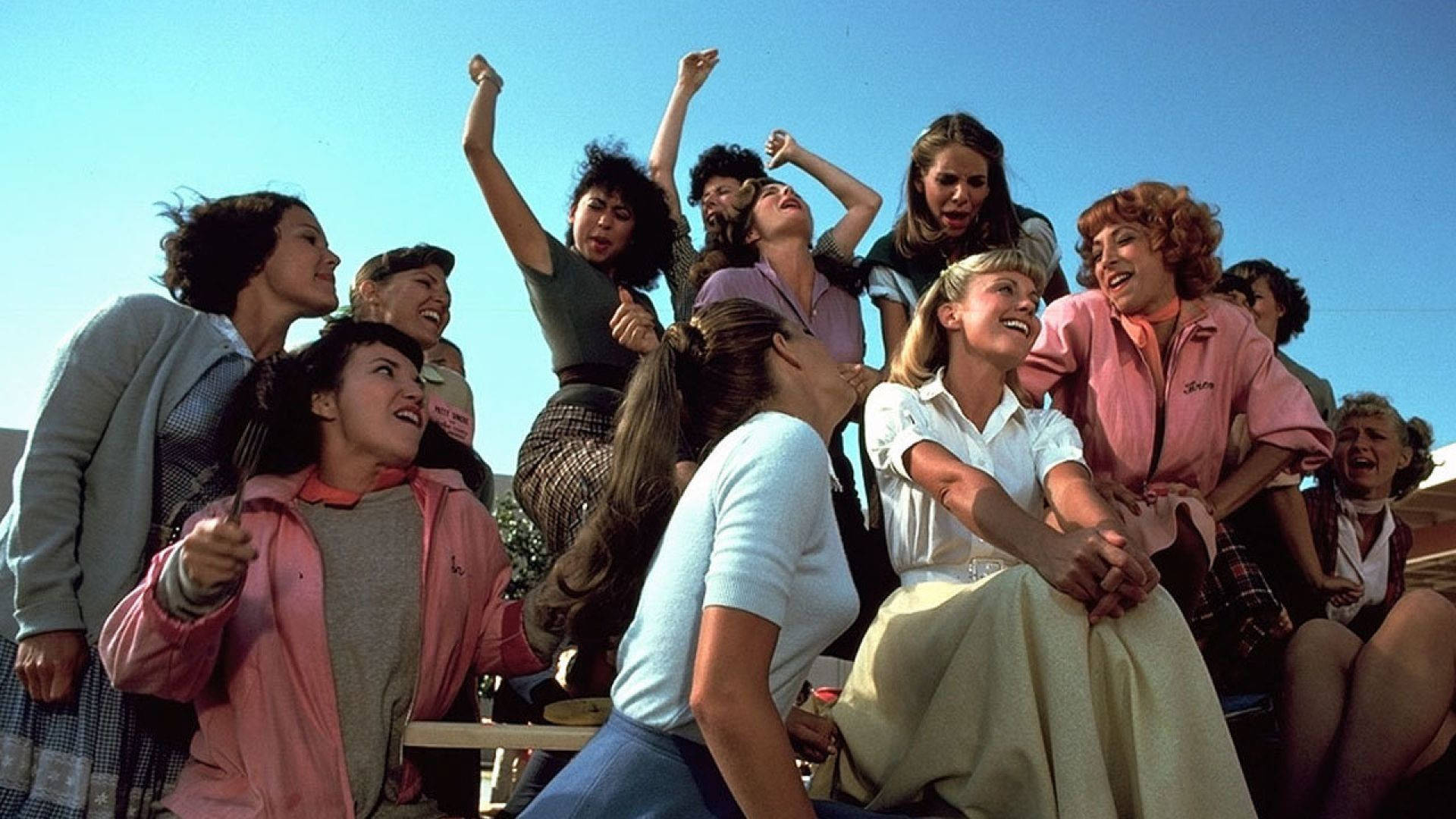 Grease 桌布 壁紙 — 一群穿著1970年代服裝的活力女性，在晴朗的藍天下舉起手臂大笑和歡呼。