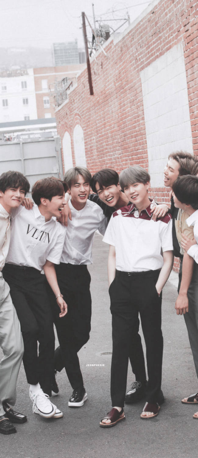 bts iPhone 桌布 壁紙 — 一群穿著休閒白襯衫和黑褲的年輕男子在陰天時靠著紅磚牆一起大笑。