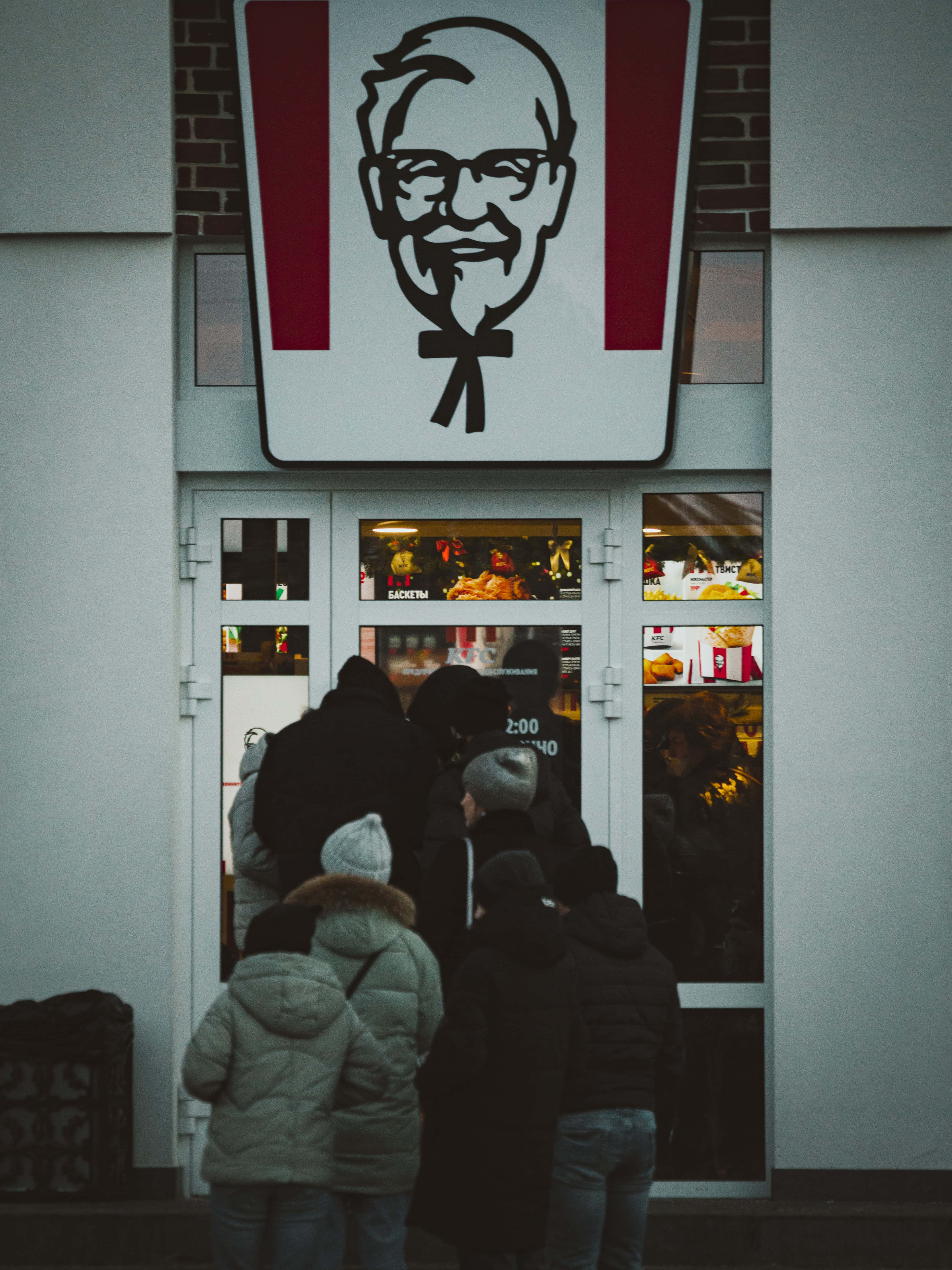 肯德基 桌布 壁紙 — 一群穿著冬季外套的人在一個大型山德士上校招牌下的KFC餐廳外面等候。