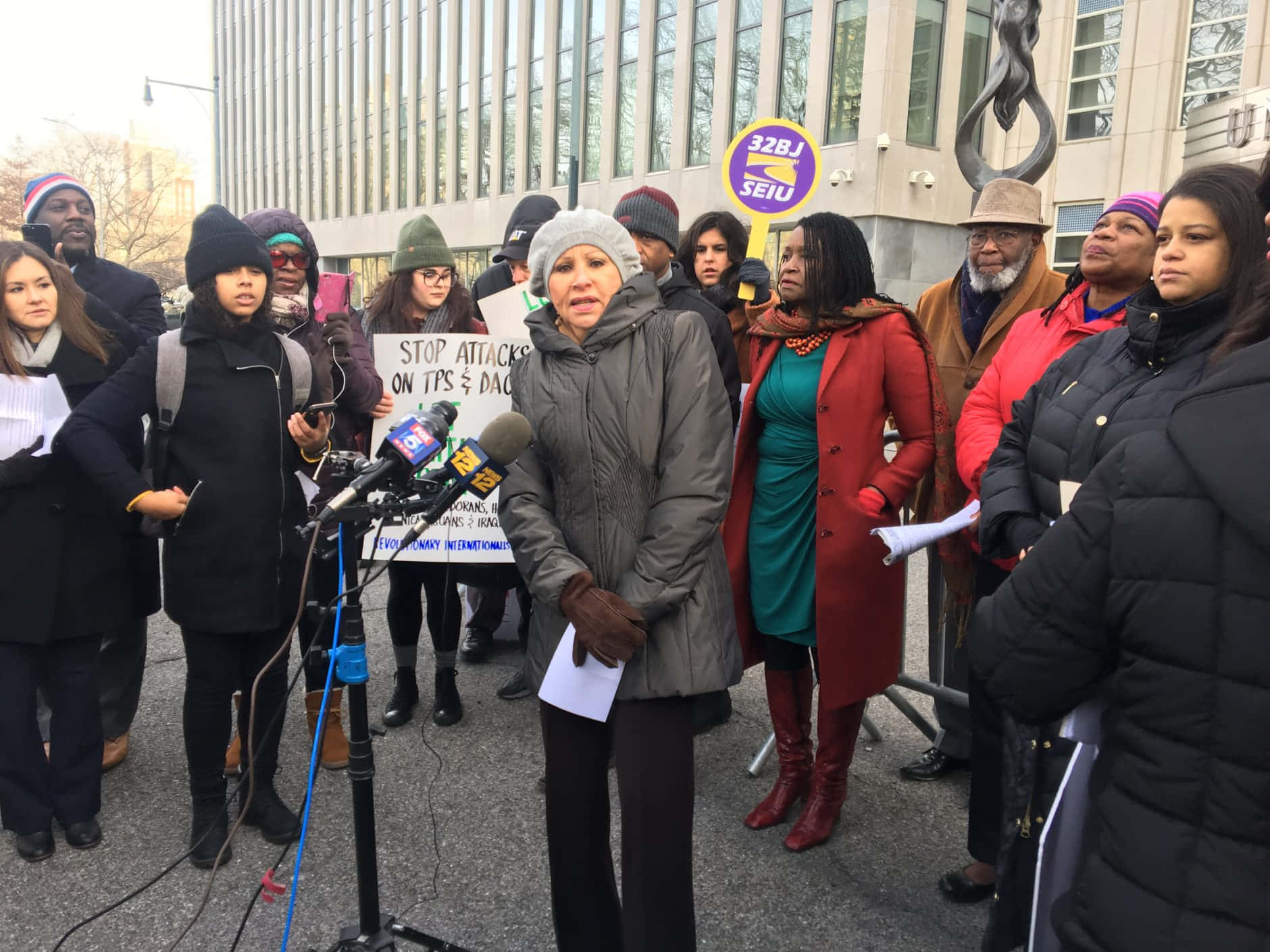 New York 桌布 壁紙 — 一群穿著冬季外套的多樣化抗議者聚集在現代建築外，手持標語並在日間集會中對著麥克風發言。