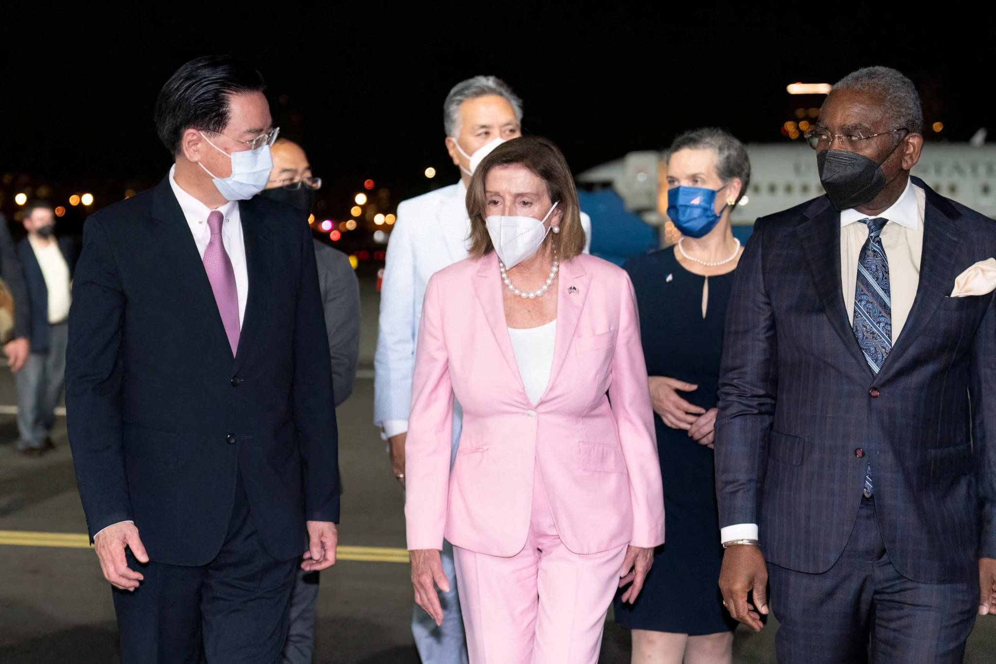 Politics 桌布 壁紙 — 一群穿著正式服裝、戴著口罩的官員在夜間行走在機場停機坪上，背景有飛機燈光閃爍。