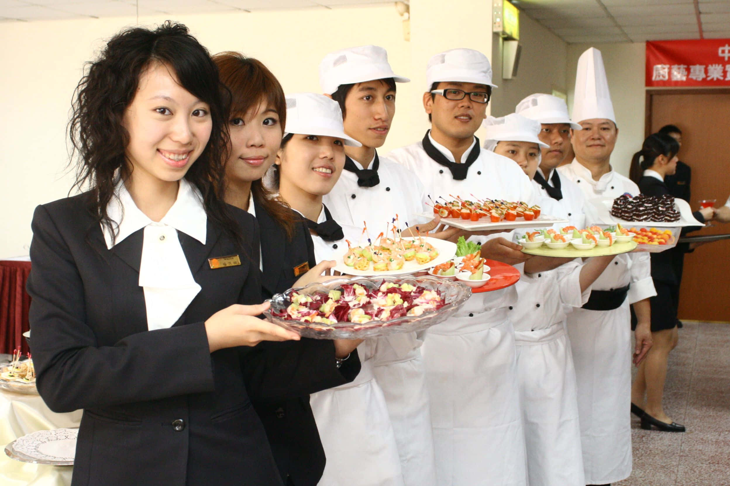 Hospitality 桌布 壁紙 — 一群穿著白色制服的愉快廚師和員工在明亮的室內廚房環境中自豪地展示色彩繽紛的盤裝開胃菜。