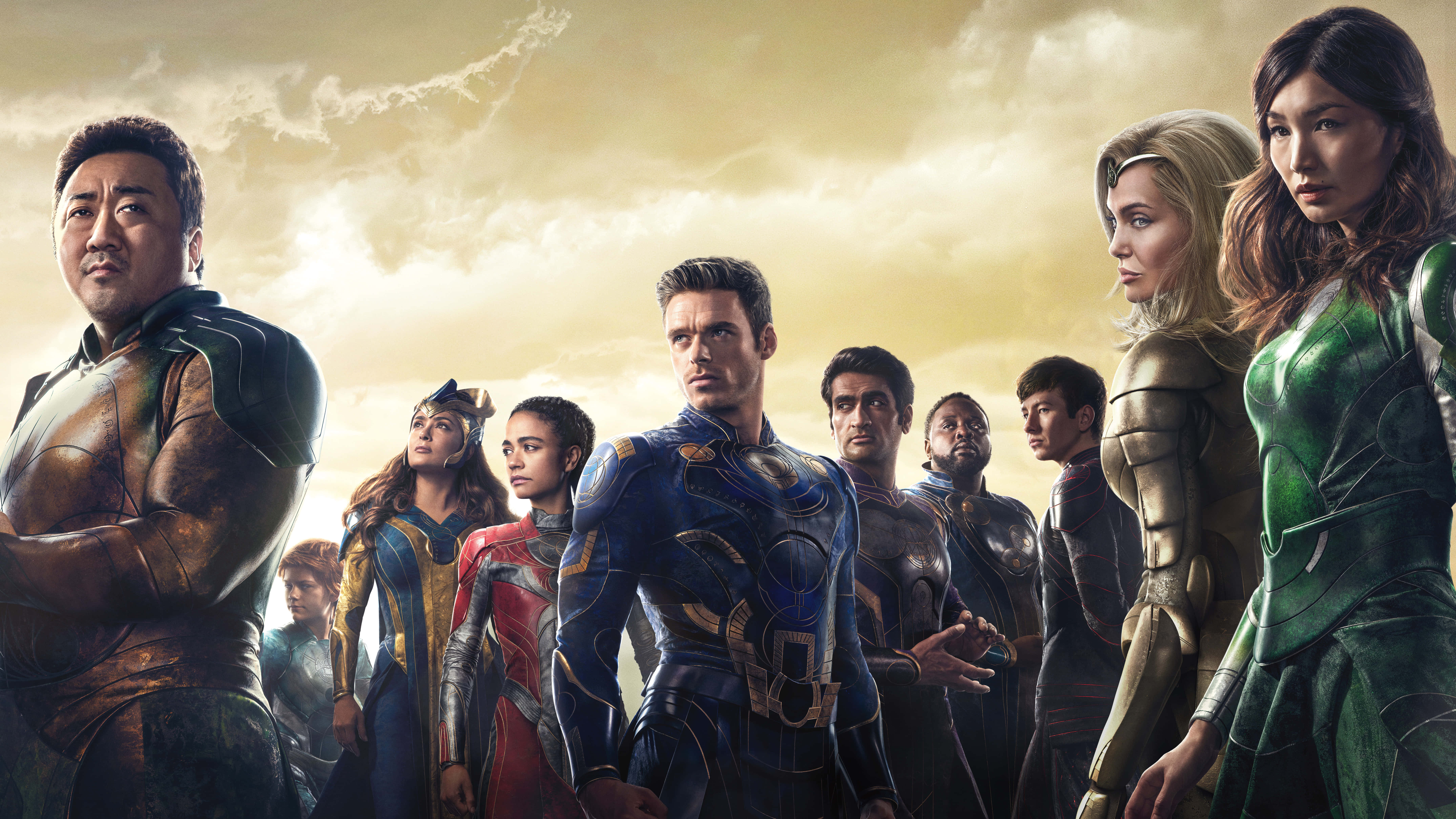 Eternals Movie 桌布 壁紙 — 一群穿著精細服裝的超級英雄自信地站在金色天空和戲劇性雲彩前。