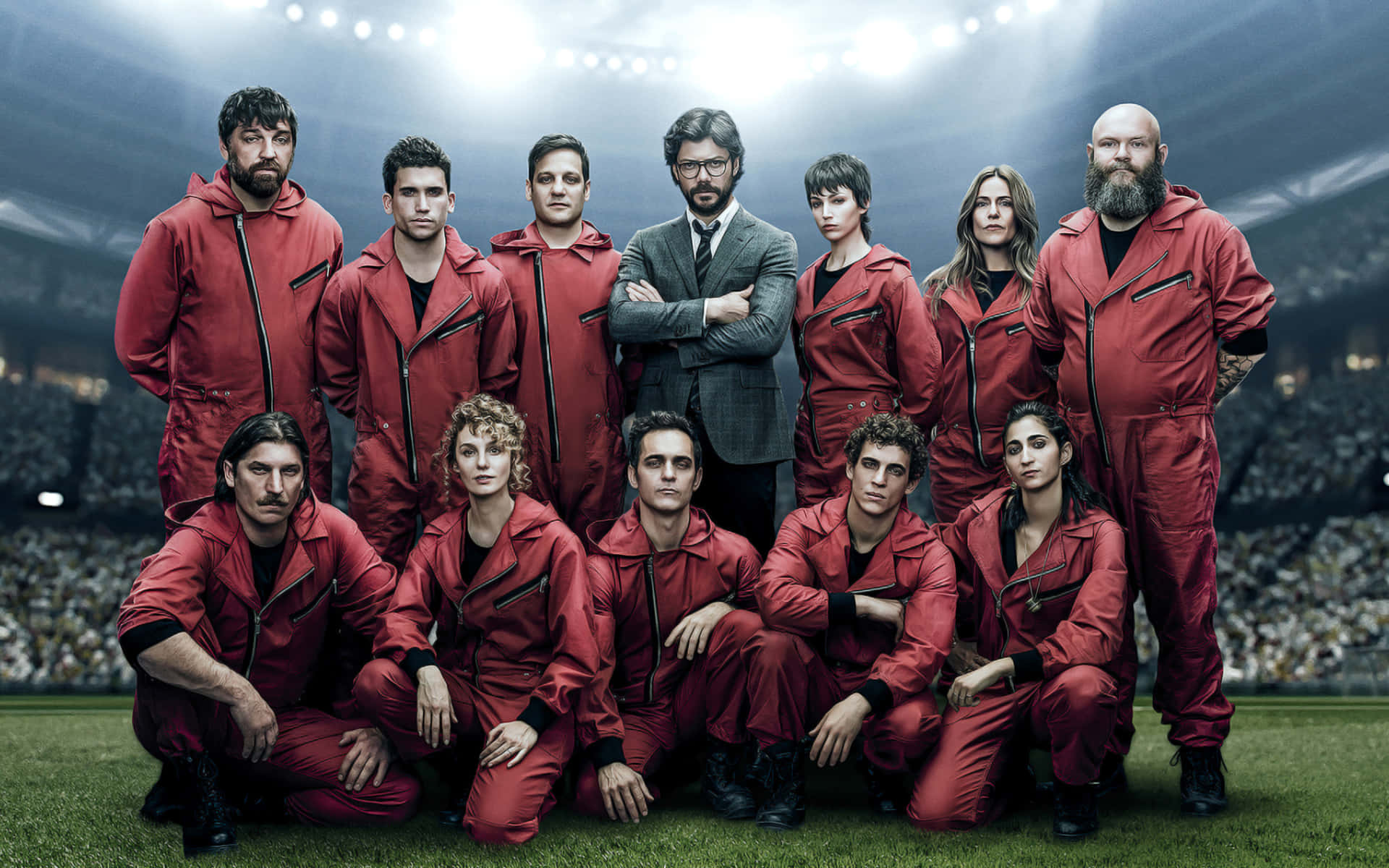 Money Heist Mask 桌布 壁紙 — 一群穿著紅色連身衣的人，在明亮的聚光燈下於體育館內擺姿勢，背景有群眾。