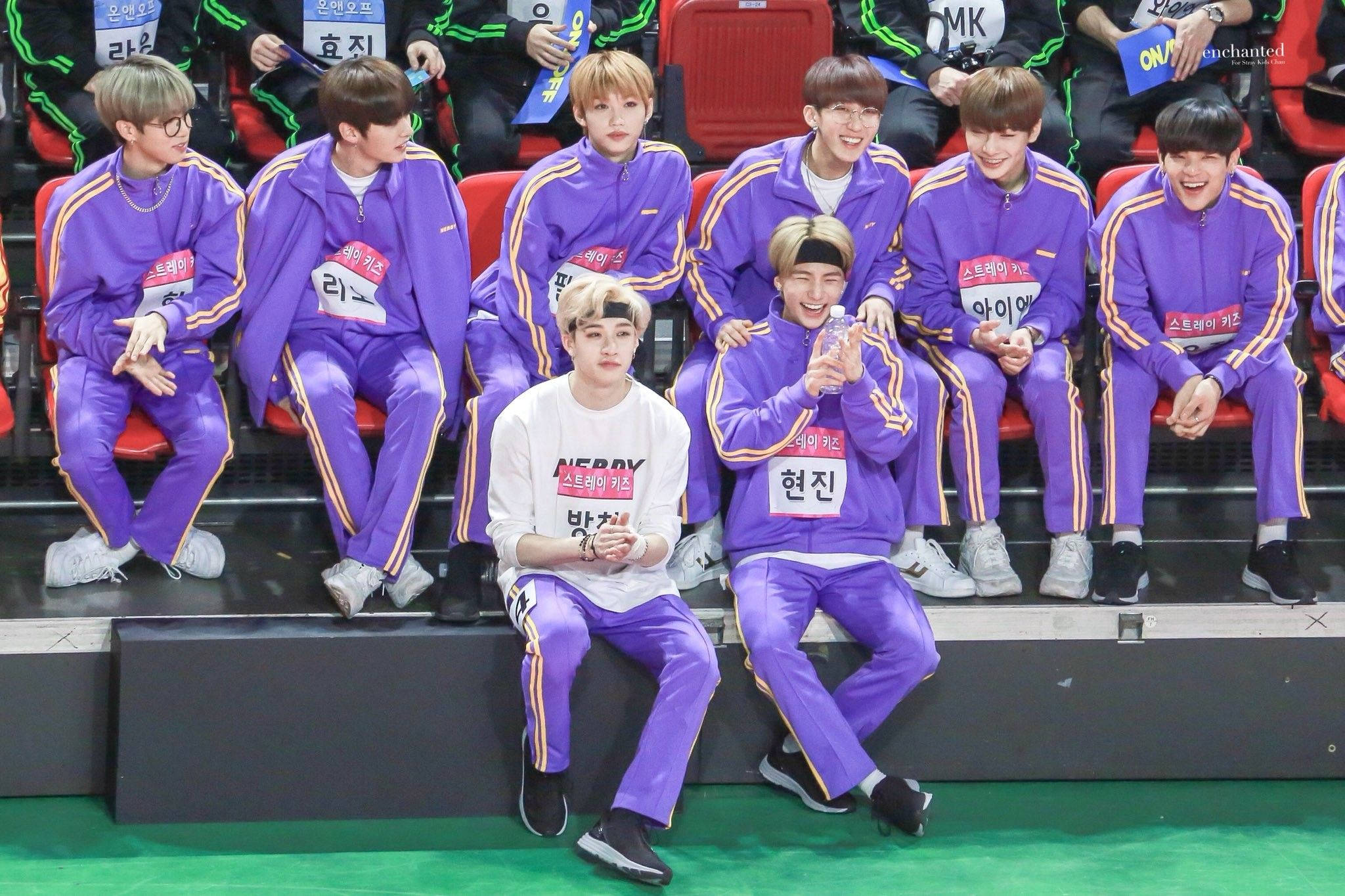 Stray Kids 桌布 壁紙 — 一群穿著紫色運動服的年輕男子在活動期間在體育館看台上大笑並擺姿勢。