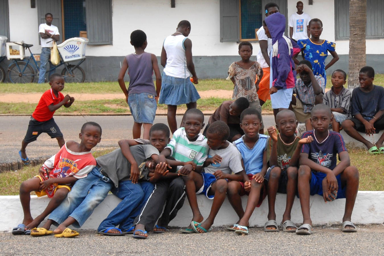Africa 桌布 壁紙 — 一群穿著色彩繽紛休閒服裝的非洲兒童，坐在和站在路邊的低矮混凝土牆上，背景有一輛送牛奶的腳踏車。