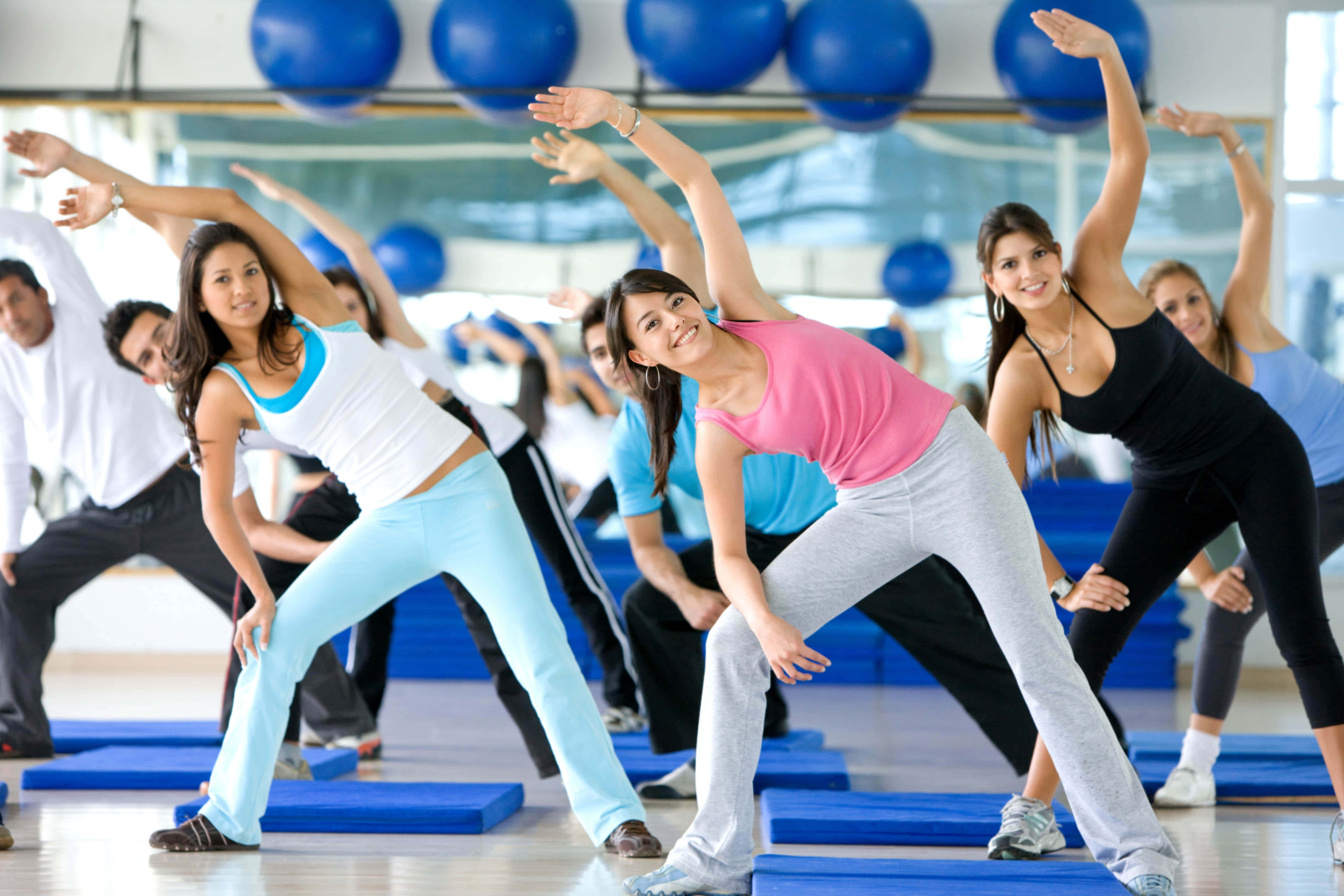 Exercise 桌布 壁紙 — 一群穿著運動服、面帶微笑的人，在明亮的健身房裡，在藍色墊子和健身球上伸展。