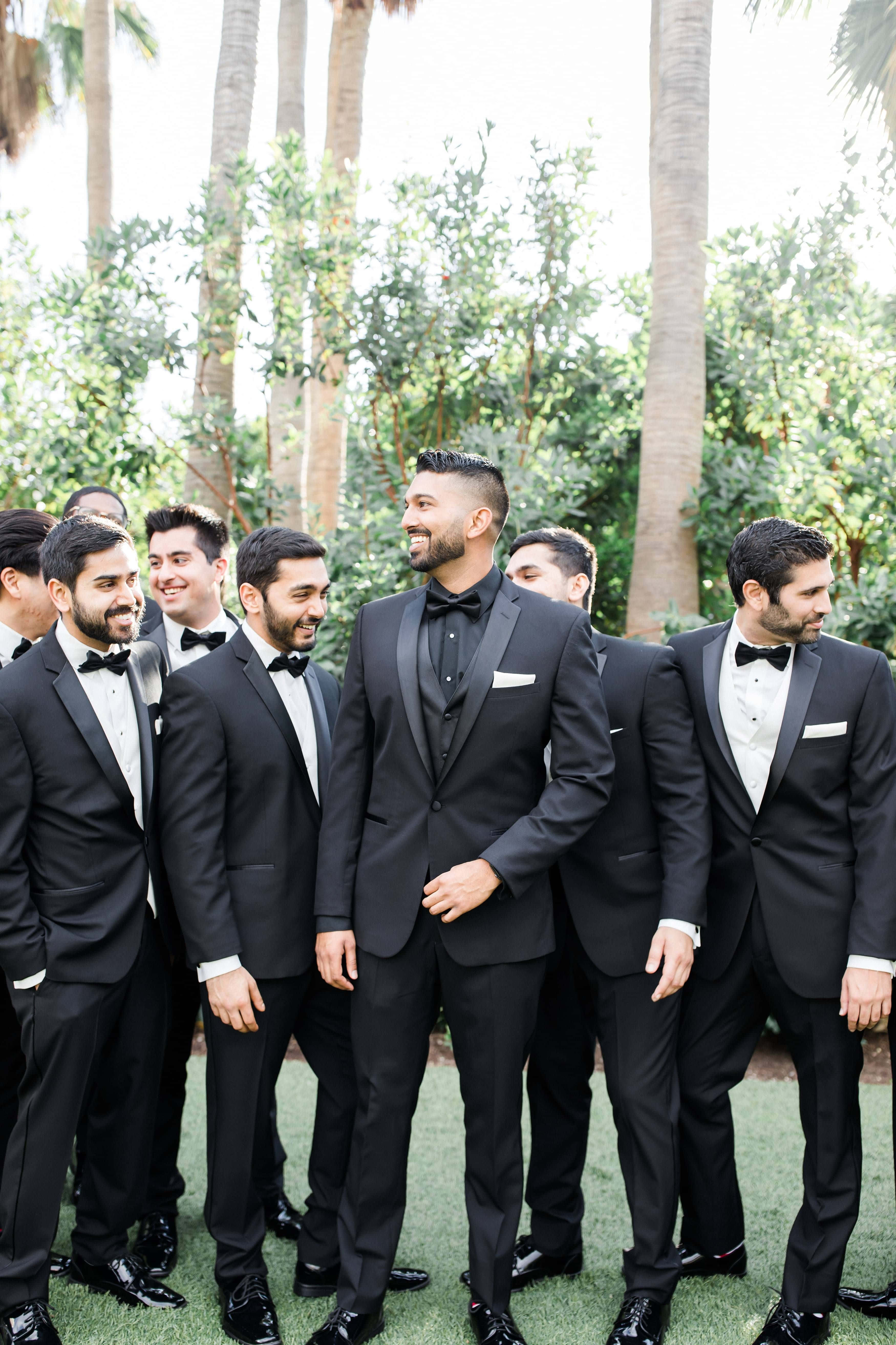 Groomsman 桌布 壁紙 — 一群穿著黑色燕尾服的伴郎在棕櫚樹下明亮的綠色草坪上一起大笑。