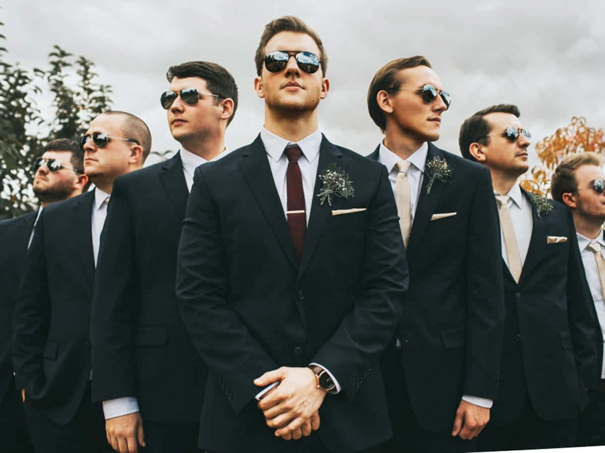 Groomsman 桌布 壁紙 — 一群穿著黑色西裝和太陽眼鏡的伴郎，在陰天背景下自信地站在戶外，背景有秋天的樹木。