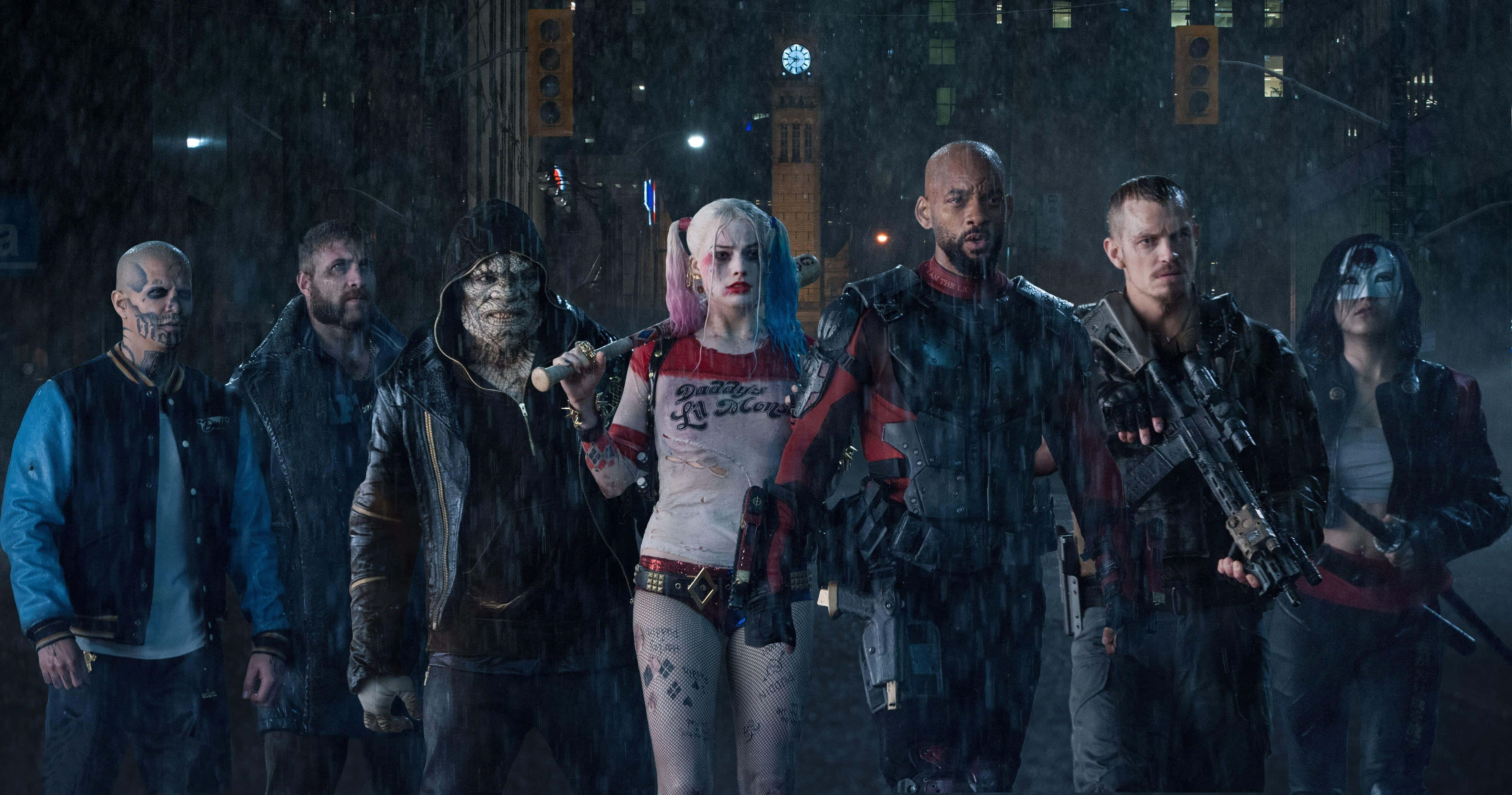 The Suicide Squad 桌布 壁紙 — 一群粗獷的武裝惡棍在夜晚站在被雨水浸透的城市街道上，頭頂是閃爍的路燈。