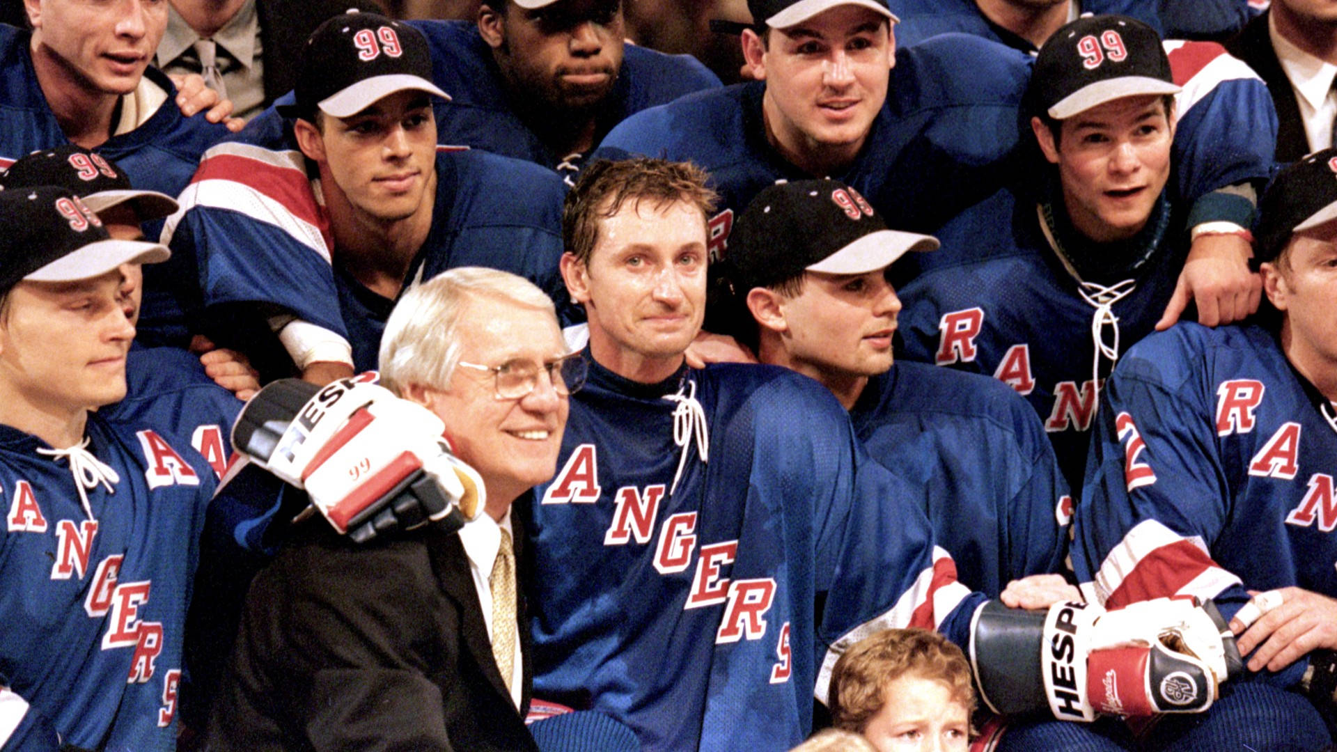 New York Rangers 桌布 壁紙 — 一群紐約遊騎兵冰球球員身穿藍色球衣、紅色字體和白色手套，與一位微笑的西裝教練合影。