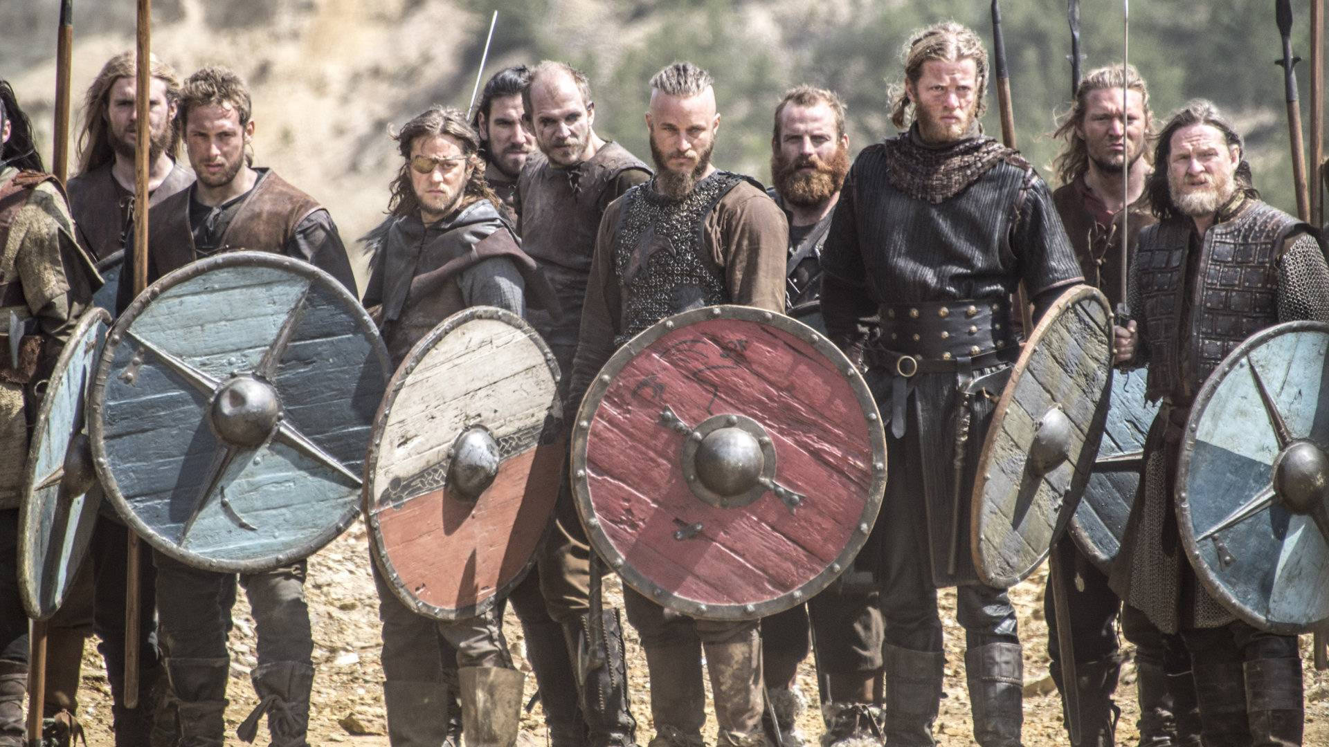 Vikings 桌布 壁紙 — 一群維京戰士排成一排，手持帶有金屬圓心的圓形木盾和長矛，站在陽光普照的岩石地形上。