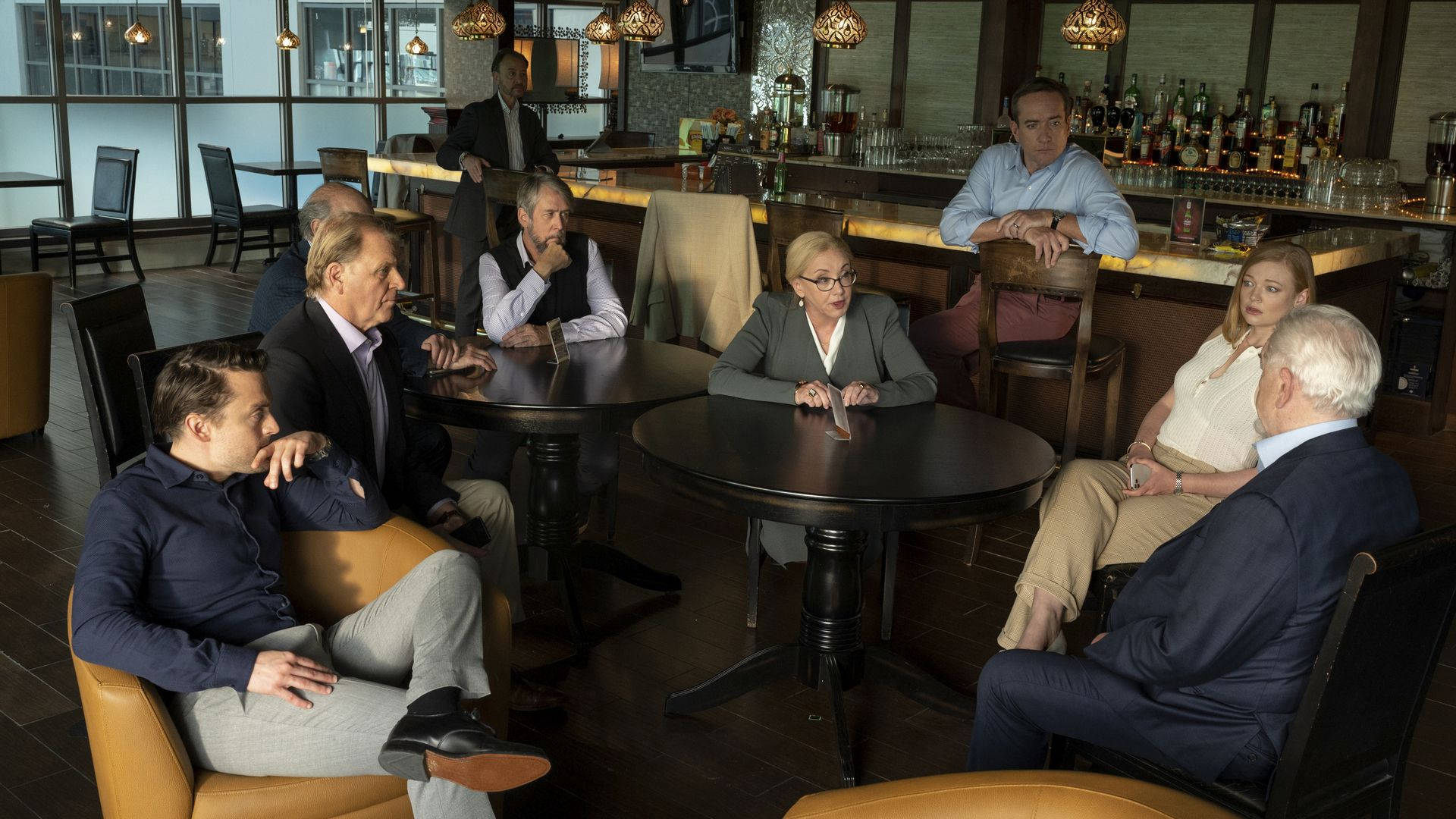 Succession 桌布 壁紙 — 一群緊張的成年人穿著商務服裝坐在現代高級酒吧的桌子旁，酒吧內有溫暖的燈光和擺滿貨品的架子。