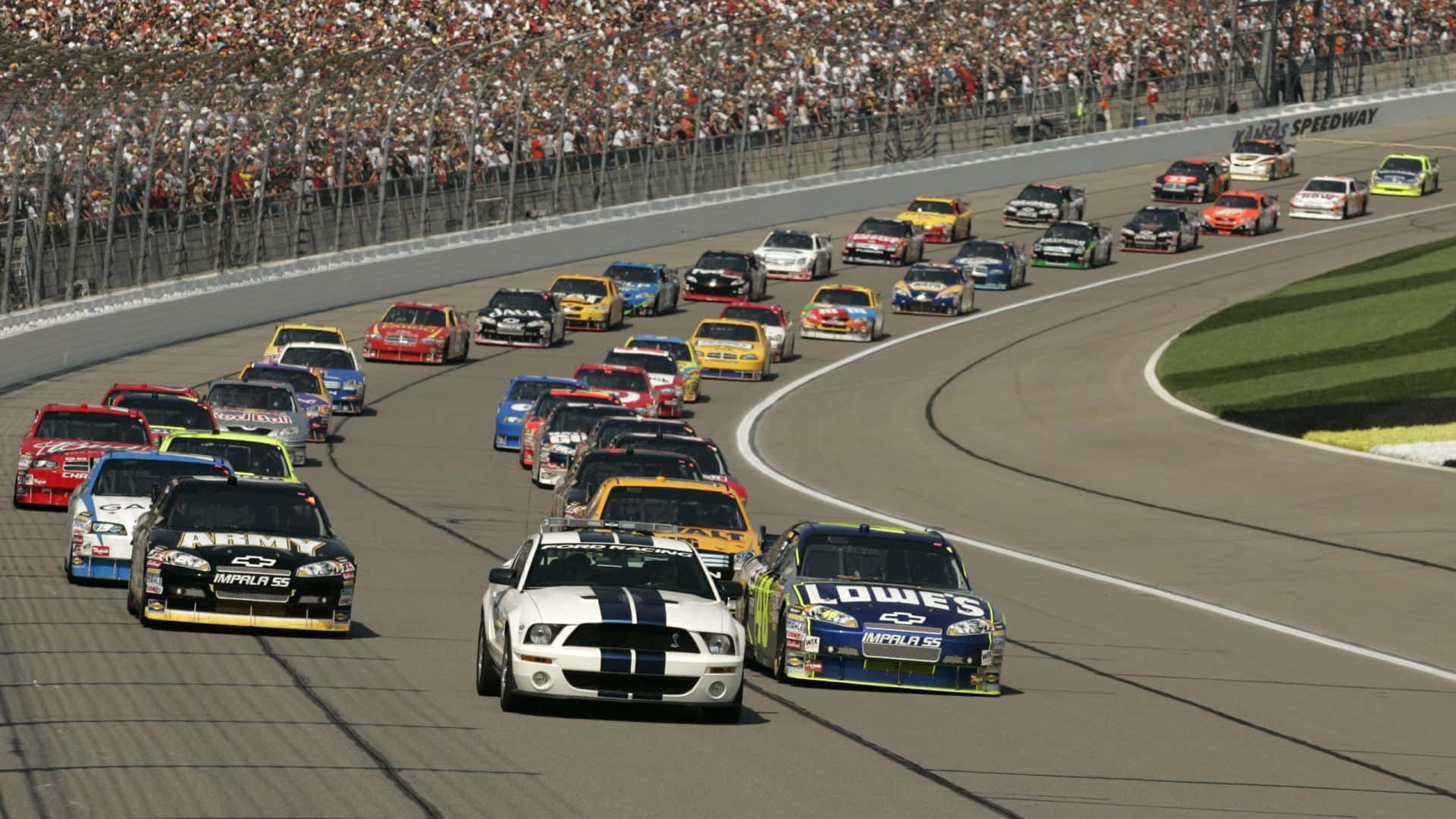 nascar 桌布 壁紙 — 一群色彩繽紛的NASCAR賽車密集地繞過陽光普照的瀝青彎道，背景是座無虛席的看台。