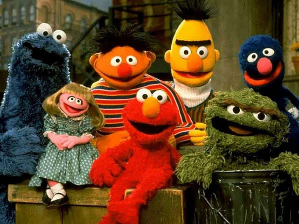 Sesame Street 桌布 壁紙 — 一群色彩繽紛的芝麻街布偶，包括 Elmo、Cookie Monster 和 Bert，一起坐在城市小巷的木箱上。