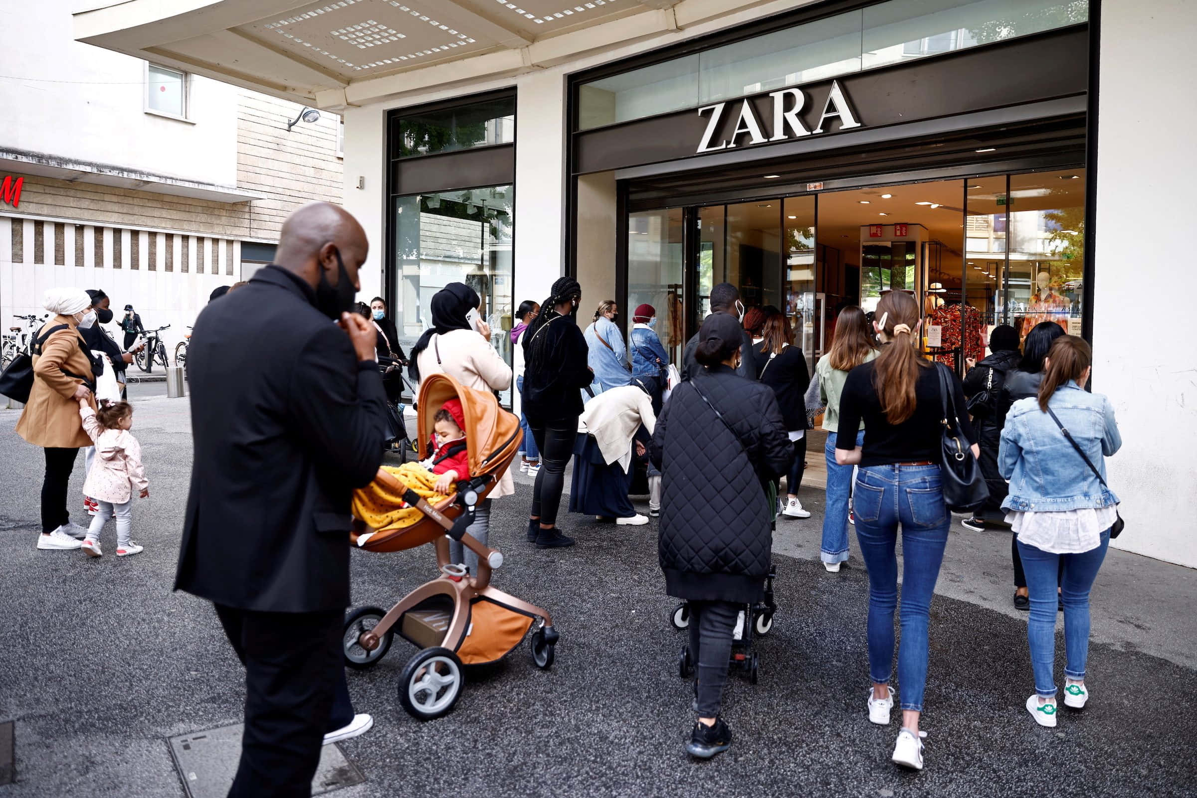 Zara 桌布 壁紙 — 一群購物者在城市人行道上，推著嬰兒車、穿著休閒服裝，在Zara商店外面排隊。
