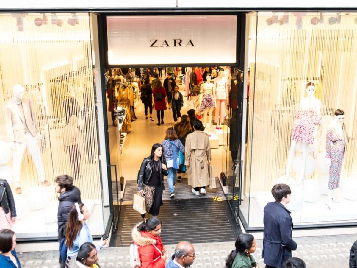 Zara 桌布 壁紙 — 一群購物者進出Zara商店，玻璃櫥窗內展示著穿著圖案服裝的人體模型。