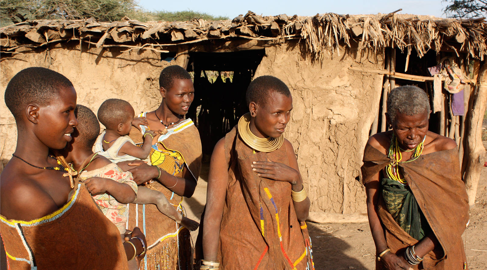 Tanzania 桌布 壁紙 — 一群身穿傳統珠飾服飾的馬賽婦女，在明亮的日光下站在茅草屋頂的泥屋前。