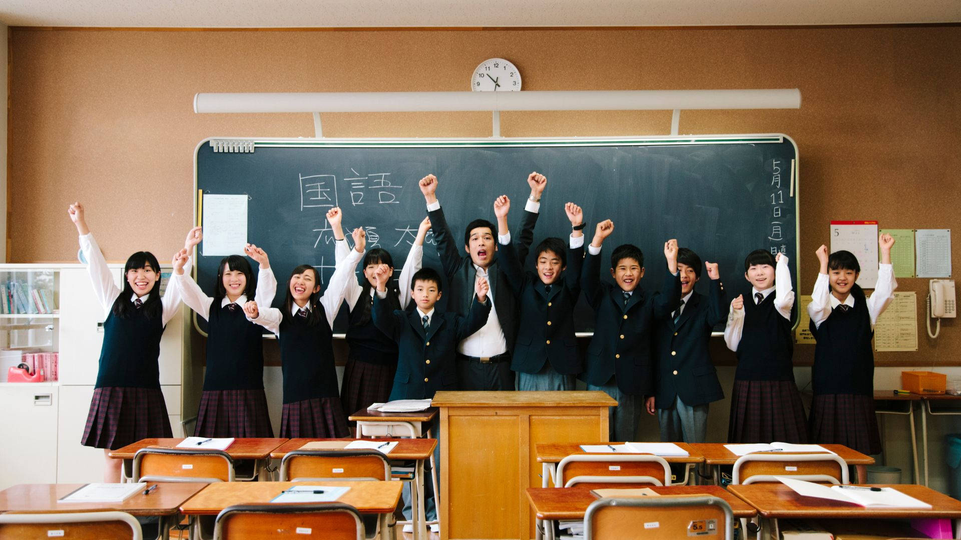 攝影 桌布 壁紙 — 一群身穿制服、歡樂的日本學童在明亮的教室裡勝利地舉起拳頭。