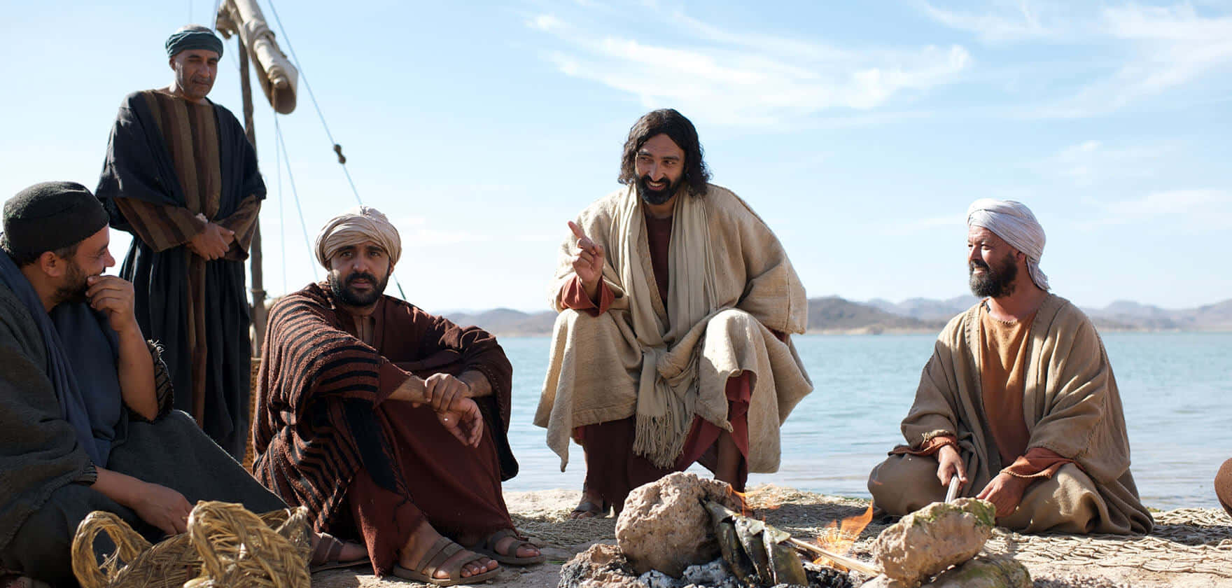 Jesus Disciples 桌布 壁紙 — 一群身穿古裝長袍的男子在晴朗藍天下，聚集在平靜湖泊旁的營火周圍。