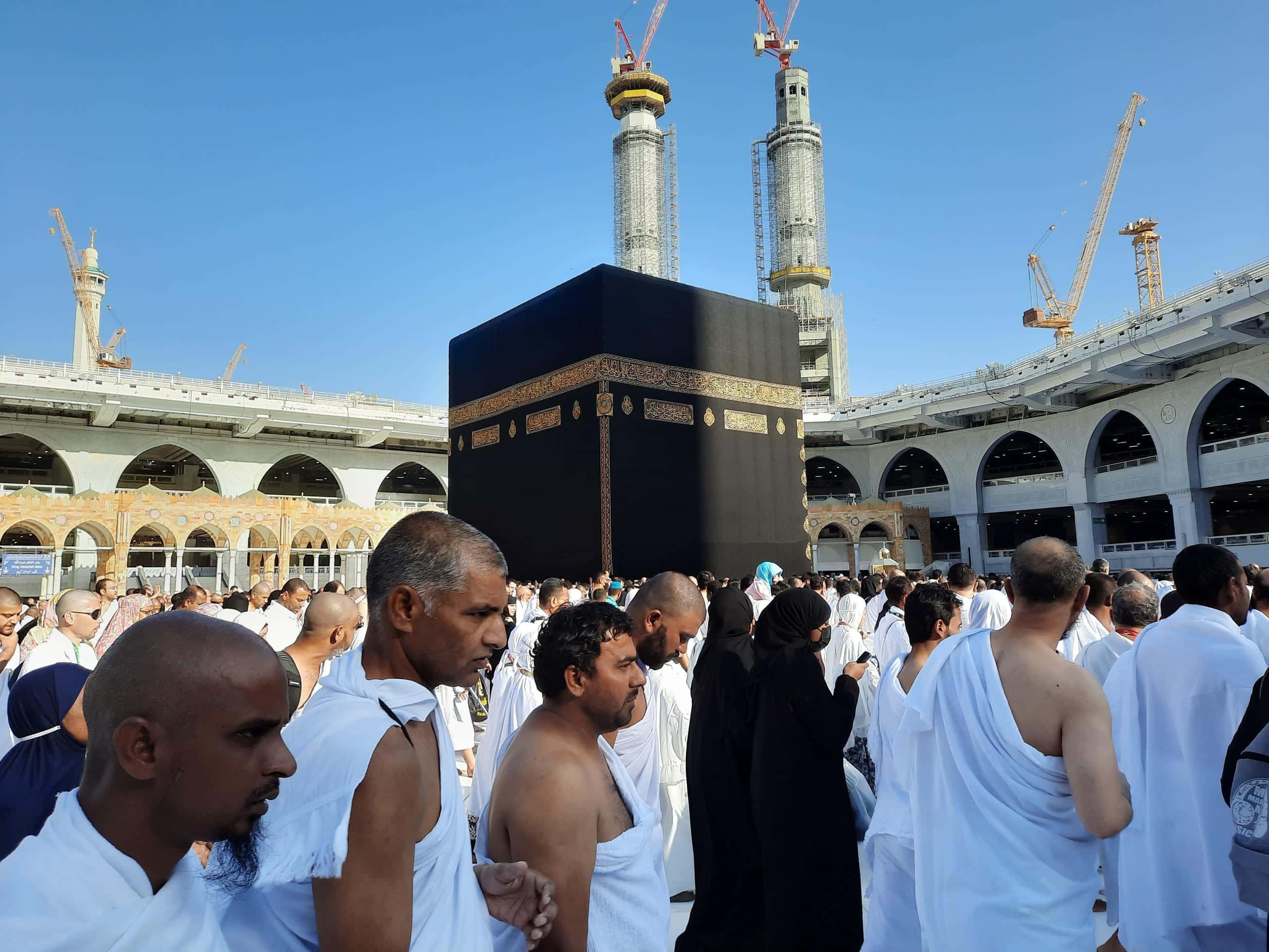 Mecca Mosque 桌布 壁紙 — 一群身穿白色朝聖服（伊赫拉姆）的朝聖者在晴朗藍天下繞行黑色天房（卡巴），建築起重機清晰可見。
