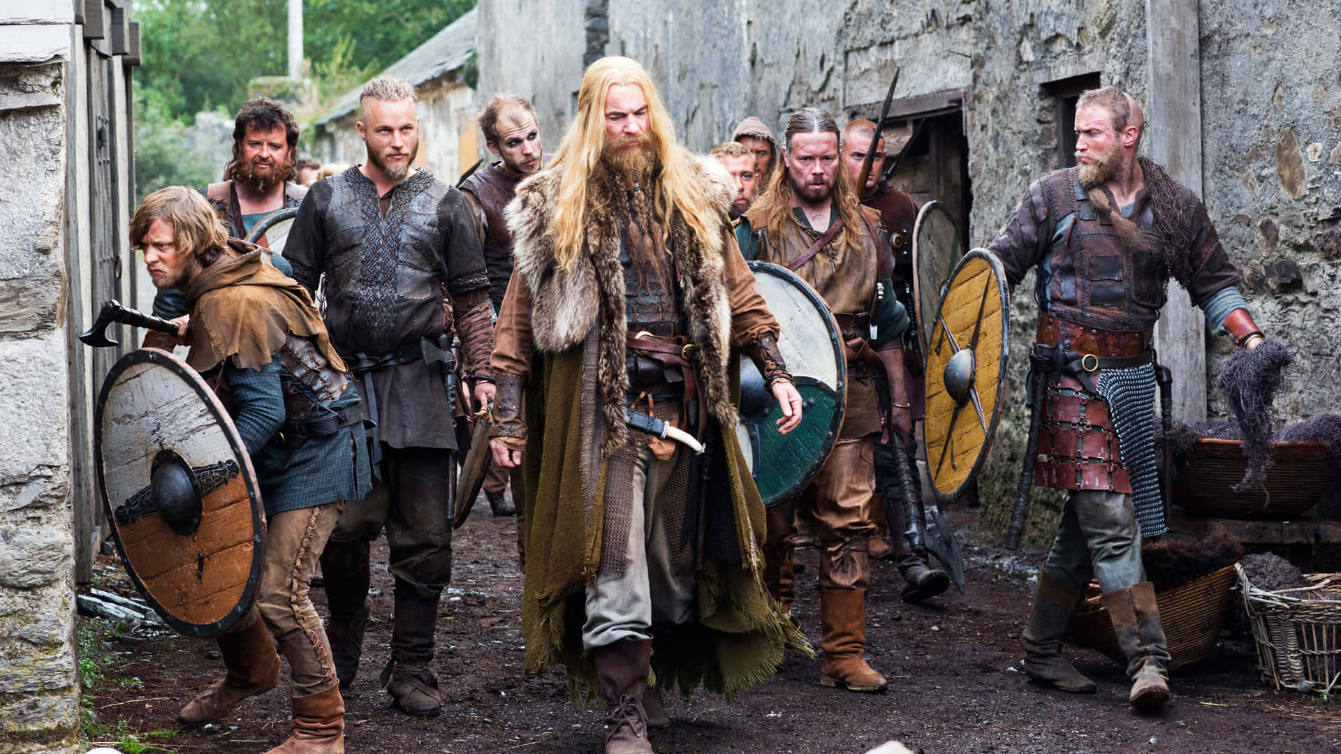 Vikings 桌布 壁紙 — 一群身穿精細盔甲和毛皮襯裡斗篷的維京戰士，手持木盾和武器，穿過石頭村莊的小巷。