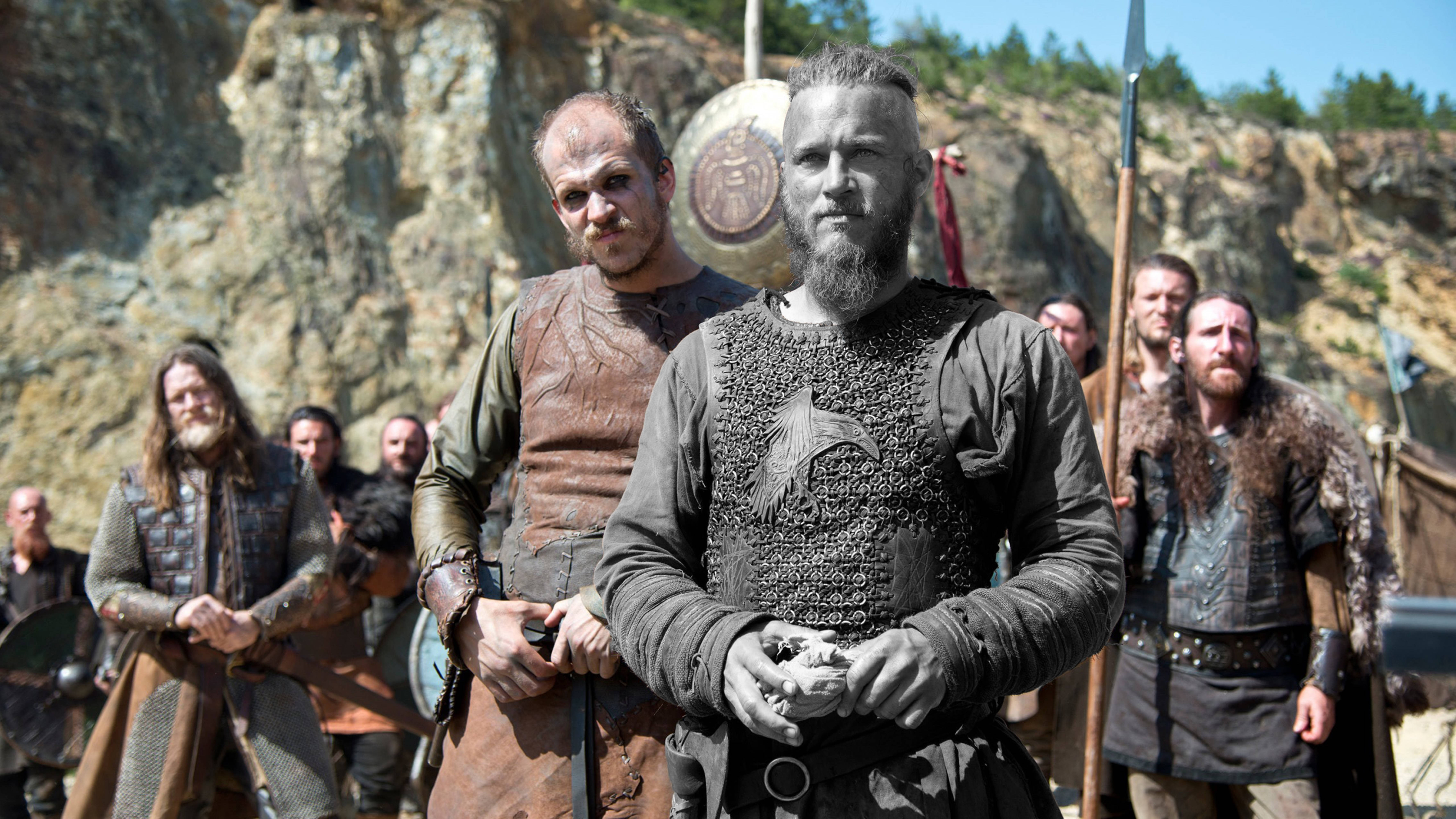 Vikings 桌布 壁紙 — 一群身穿精細盔甲的維京戰士在明亮的日光下站在岩石峭壁前。