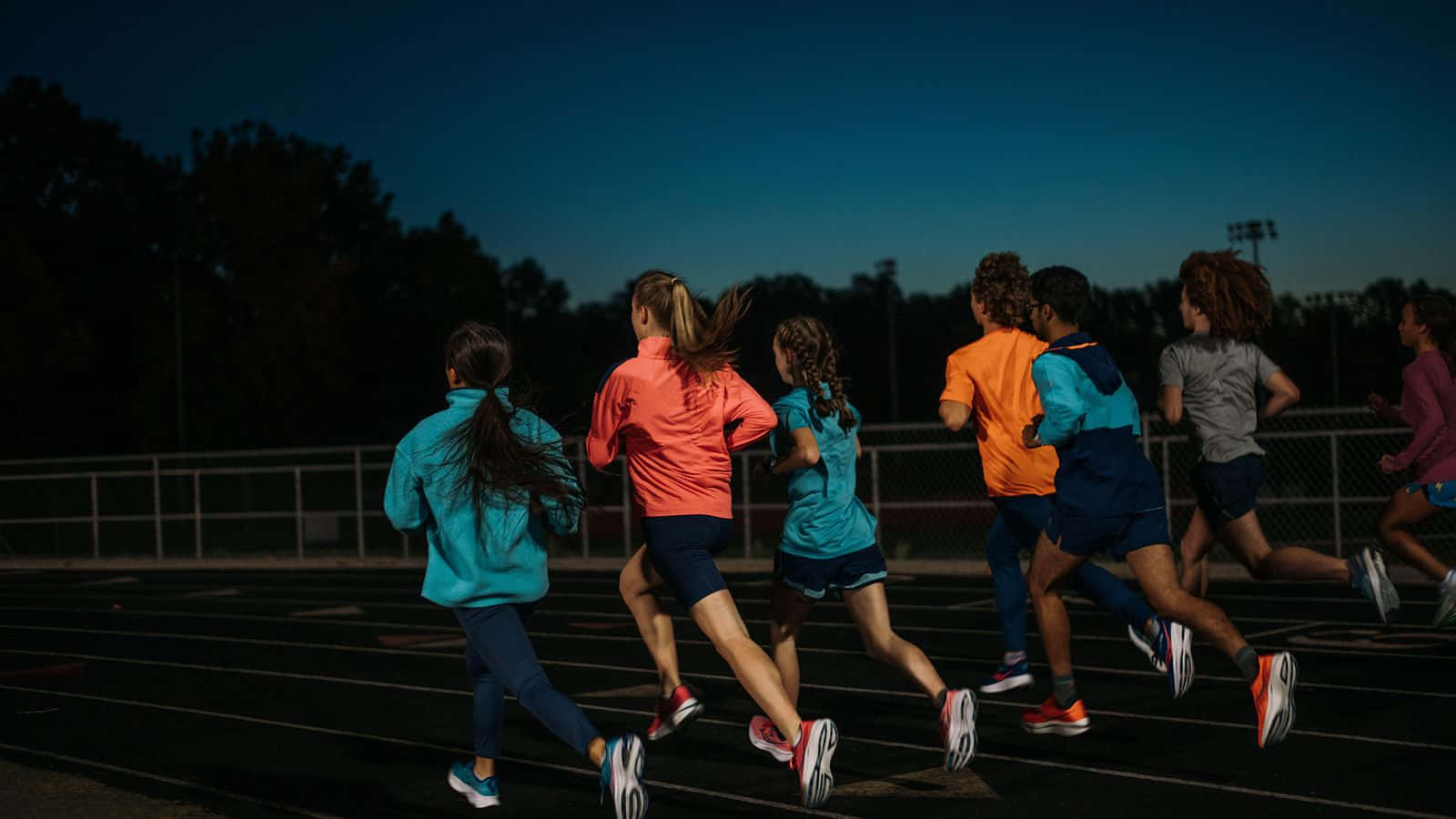 Running 桌布 壁紙 — 一群身穿色彩繽紛運動服的跑者在暮色中的跑道上衝刺，背景有樹木和圍欄。