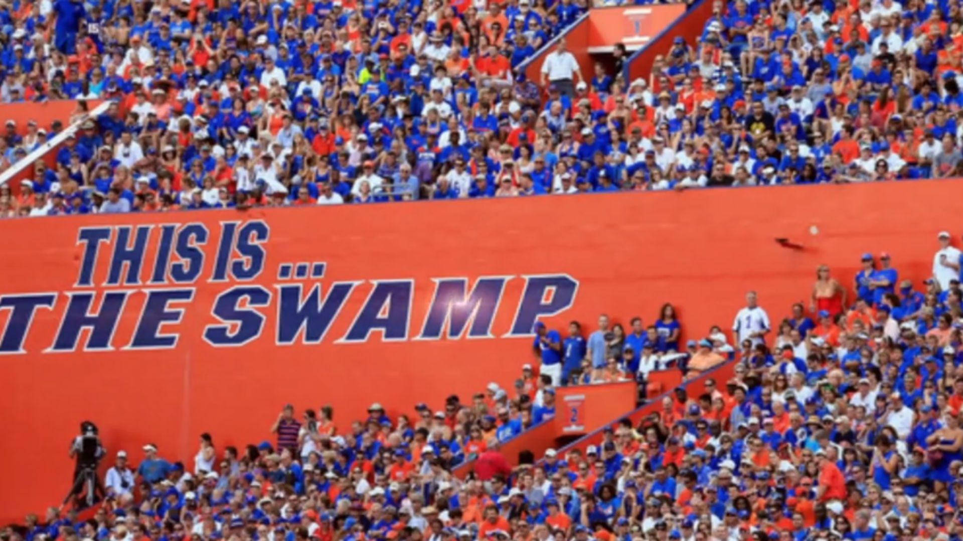 Florida Gators 桌布 壁紙 — 一群身穿藍色和橙色衣服的球迷在一個有巨大橙色牆壁、上面寫著 'THIS IS... THE SWAMP' 的體育場內歡呼。