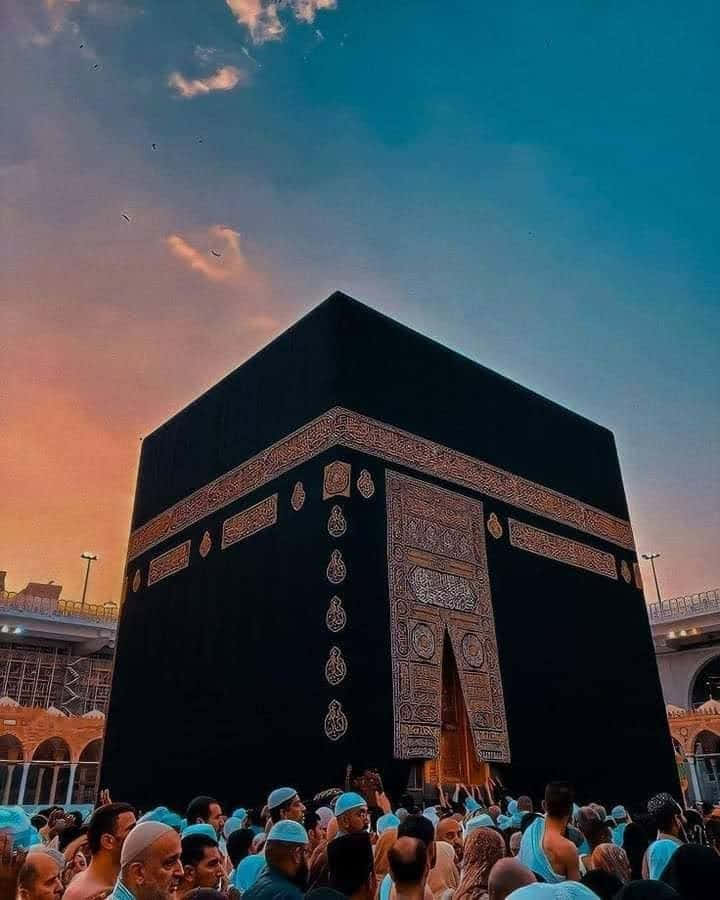 Mecca Kaaba 桌布 壁紙 — 一群身穿藍色朝聖服的朝聖者在日落時分圍繞著黑色卡巴天房，天空呈現鮮豔的橙色和藍色。