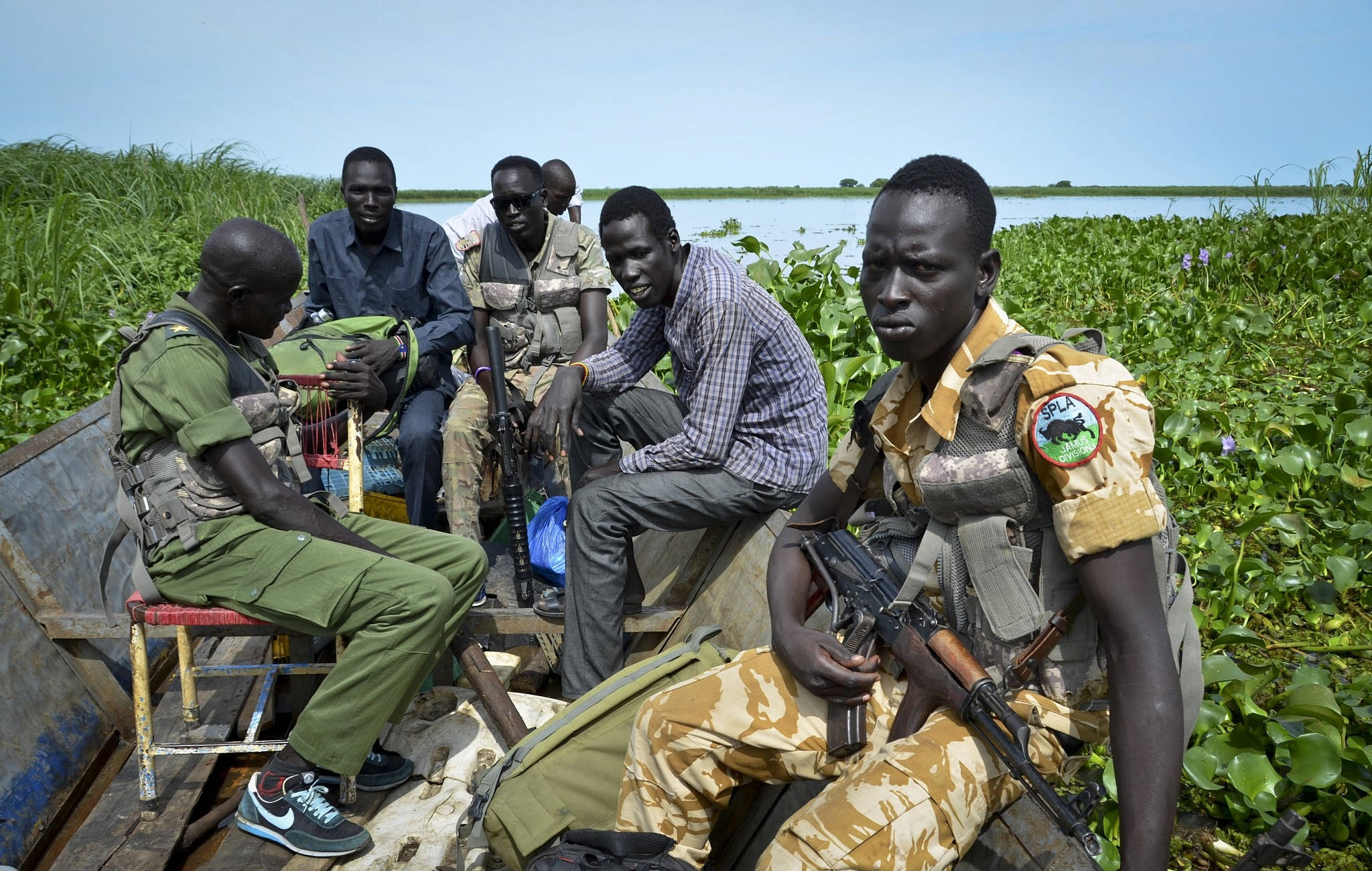 South Sudan 桌布 壁紙 — 一群身穿軍裝的武裝男子坐在木船上，周圍是茂密的綠色水生植物，頭頂是清澈的藍天。