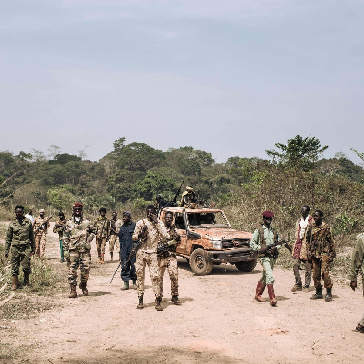 African 桌布 壁紙 — 一群身穿迷彩服的武裝士兵站在乾燥稀樹草原景觀中的泥路上，圍繞著一輛生鏽的軍用卡車。