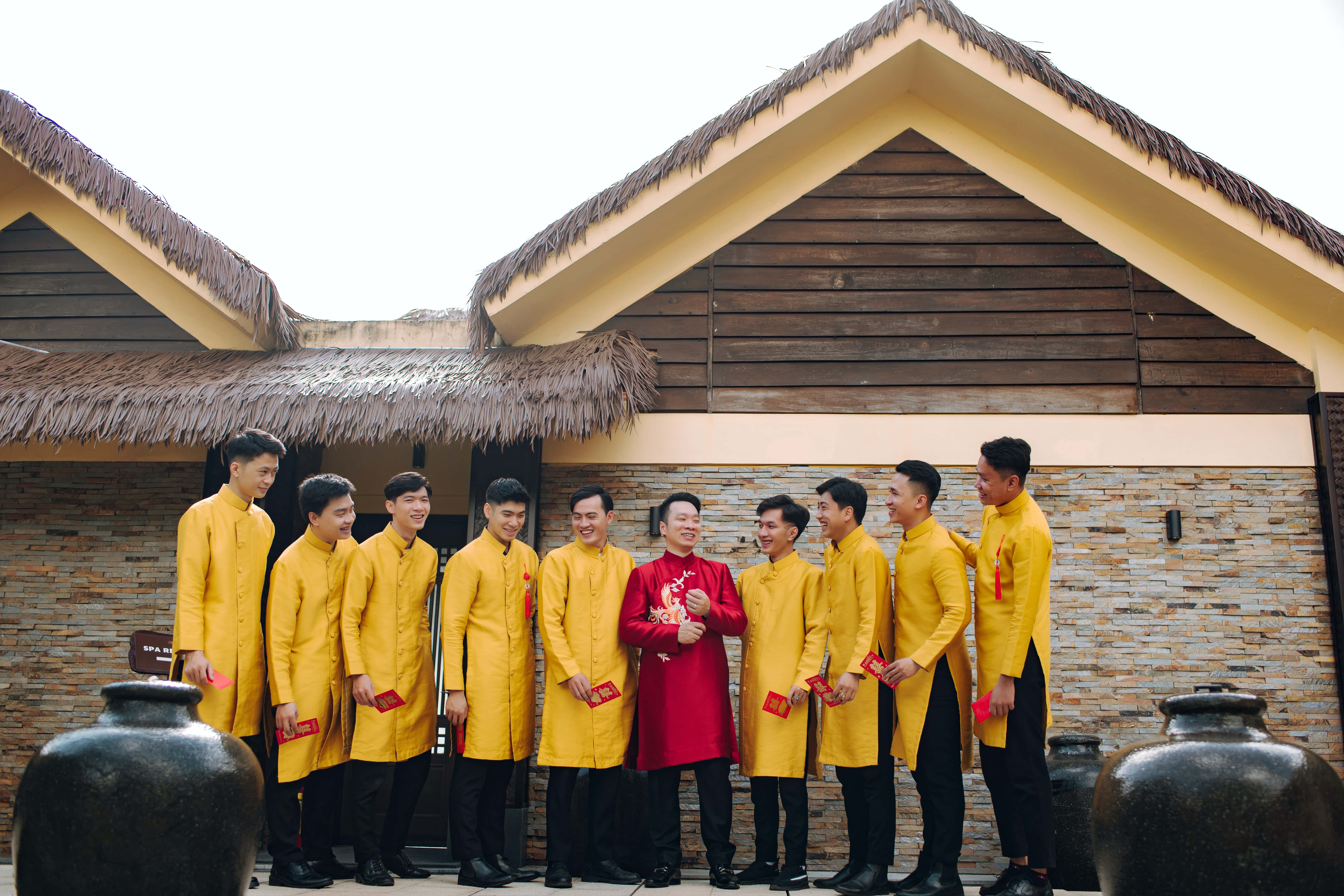 Groomsman 桌布 壁紙 — 一群身穿鮮黃色傳統服飾的男子，在茅草屋頂石牆建築前微笑站立。