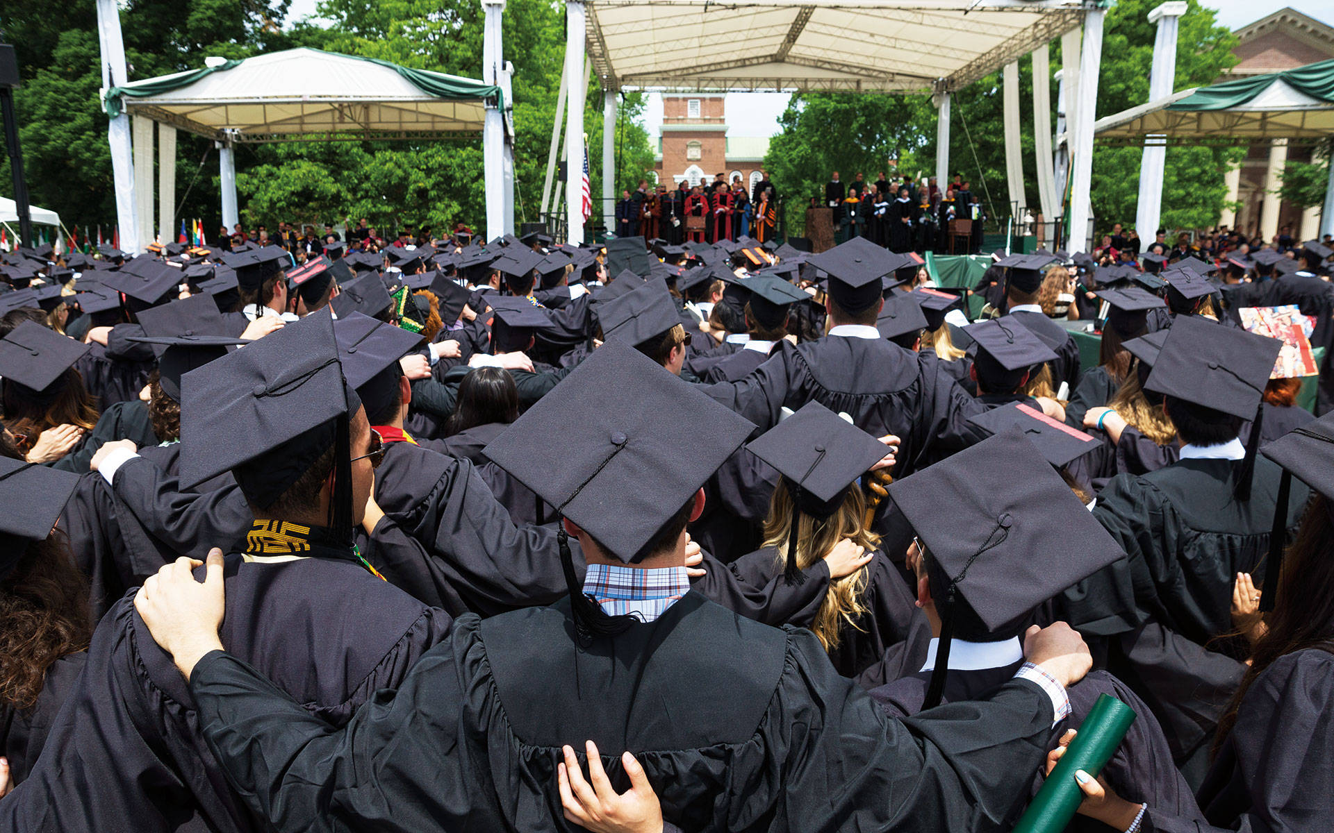 Graduation 桌布 壁紙 — 一群身穿黑色學士帽和畢業袍的畢業生在陽光明媚的戶外畢業典禮上，在白色帳篷下緊緊擁抱。