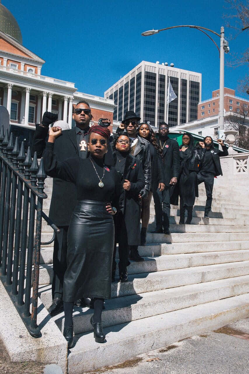 Black Panther Party 桌布 壁紙 — 一群身穿黑衣的黑人活動家，在晴朗的藍天下，有力地站在歷史建築前的石階上。