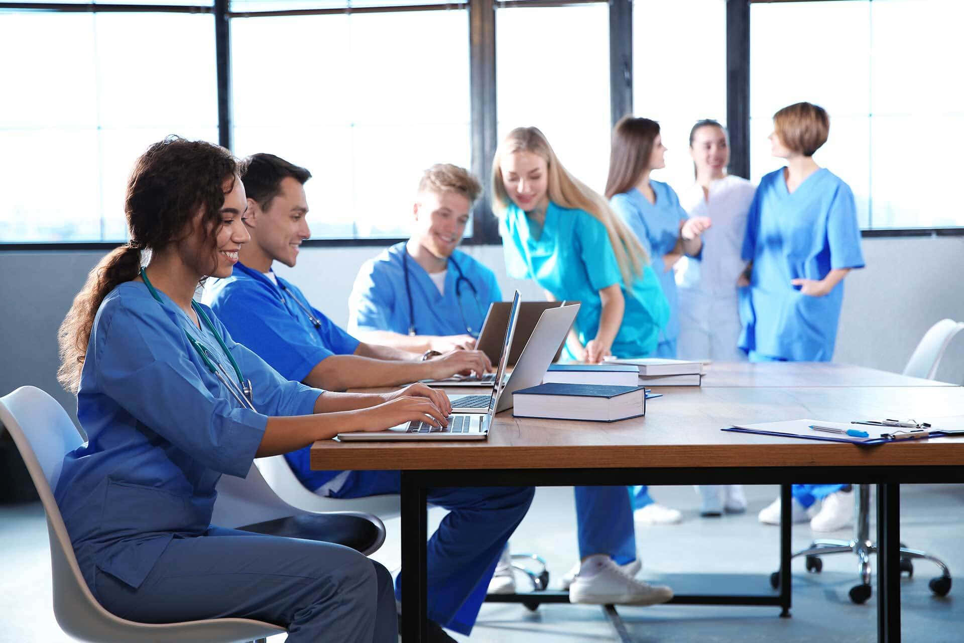 Nursing 桌布 壁紙 — 一群面帶微笑的醫護專業人員穿著藍色手術衣，在明亮的現代辦公室裡圍著木桌，使用筆記型電腦和書籍進行協作。