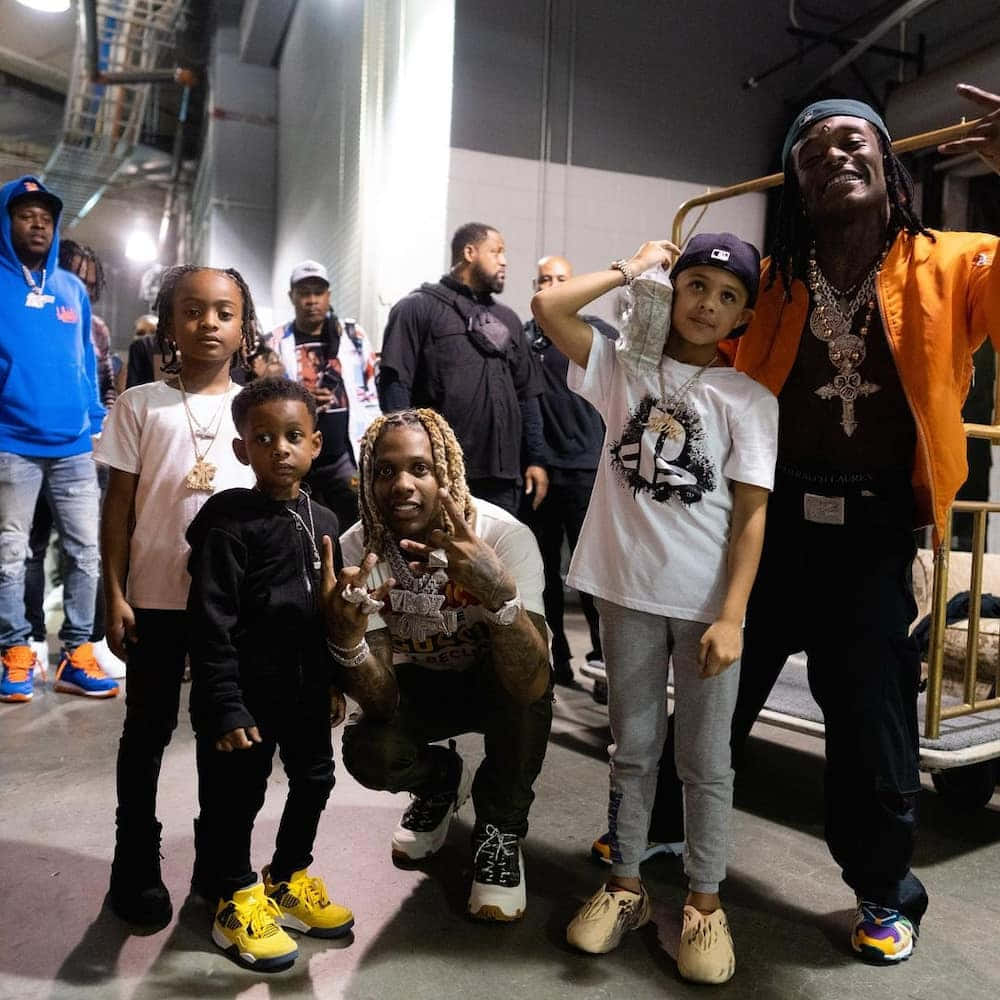 Lil Durk 桌布 壁紙 — 一群饒舌歌手和兒童在昏暗的後台區域，在工業照明和金屬結構下一起擺姿勢。