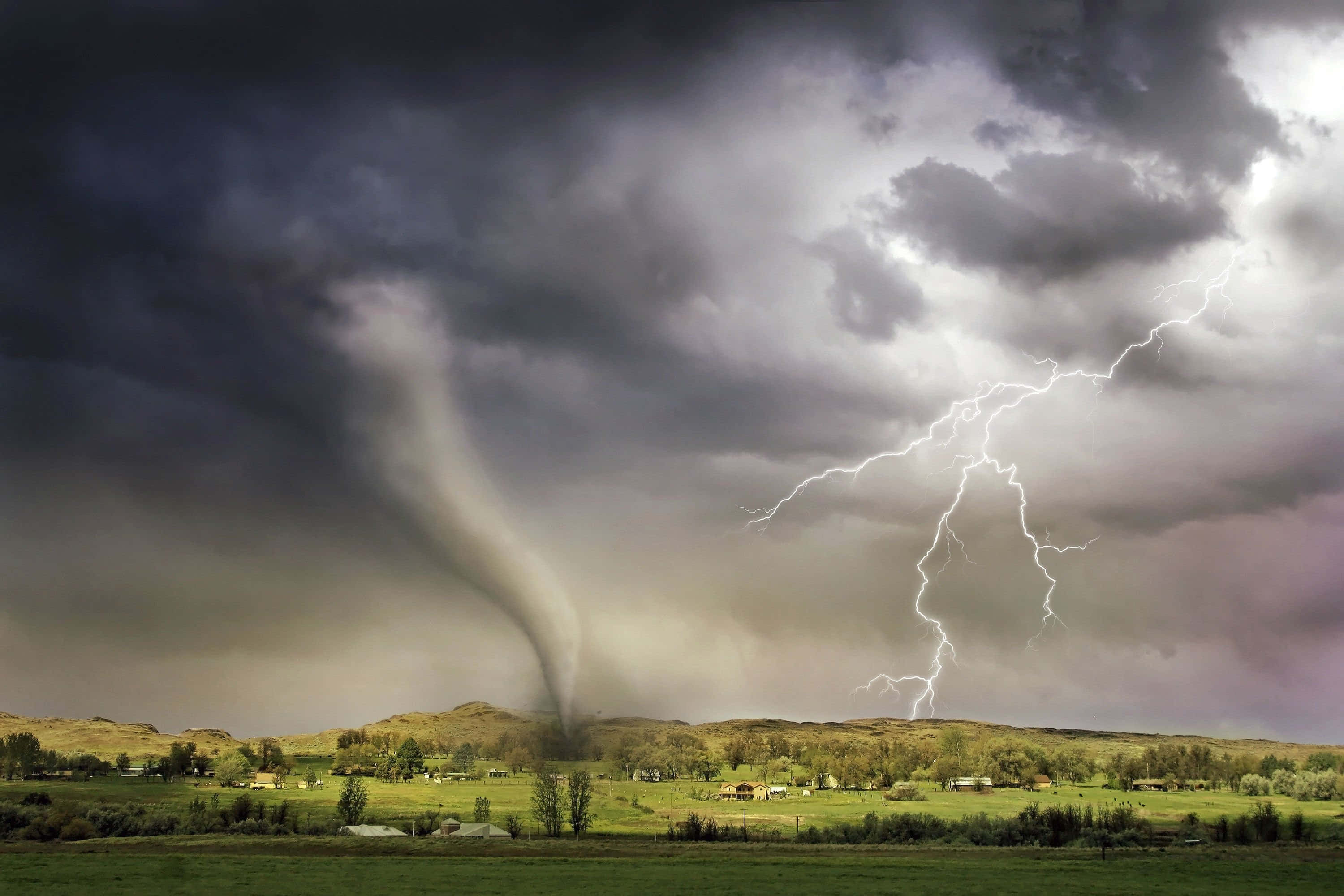 暴風 桌布 壁紙 — 一股強大的龍捲風扭曲穿過鄉村景觀，閃電擊中散落房屋上方暴風雨的天空。
