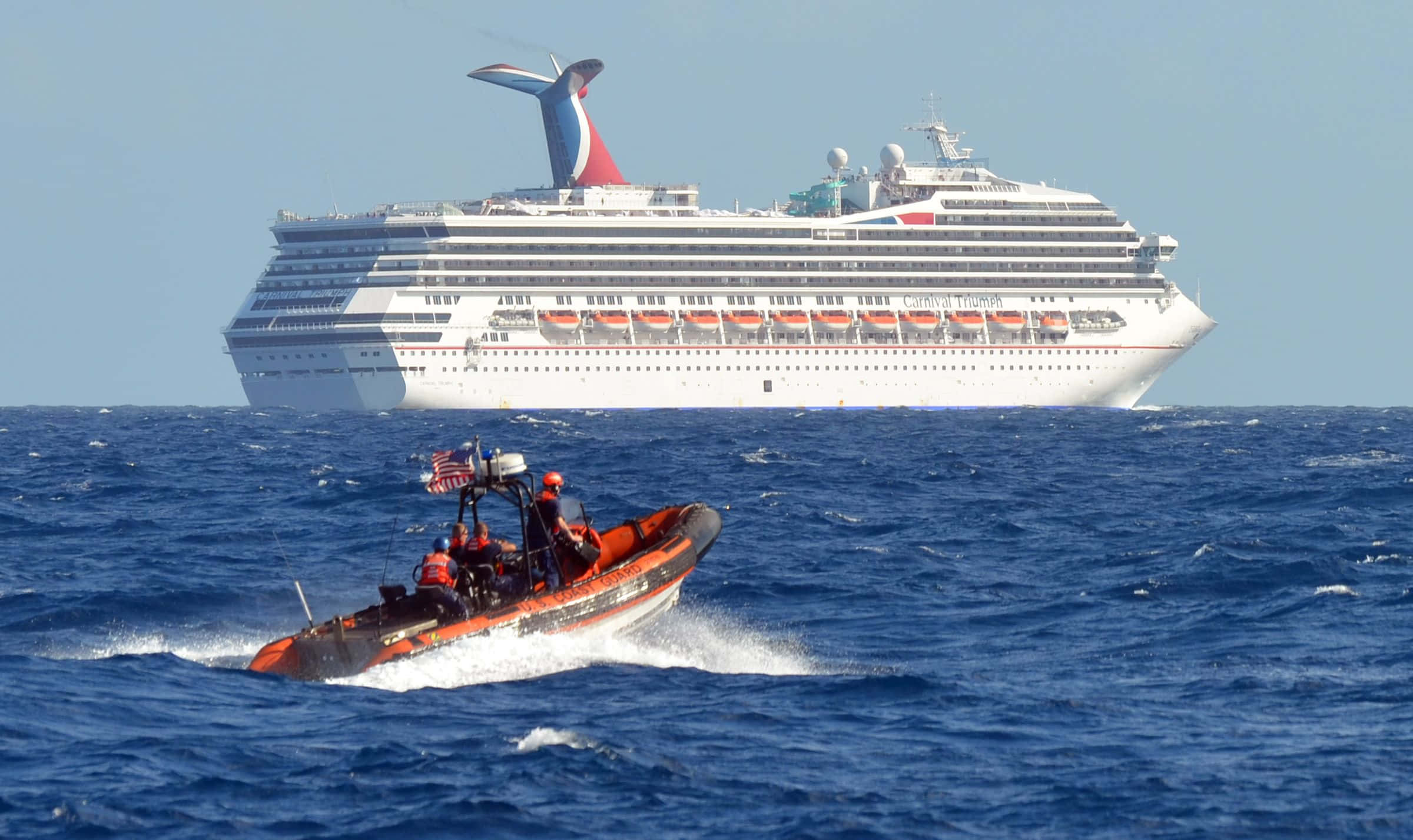Carnival Dream 桌布 壁紙 — 一艘亮橙色的海岸防衛隊救援艇在波濤洶湧的藍色海面上飛馳，背景中一艘巨大的白色嘉年華郵輪在晴朗的天空下若隱若現。