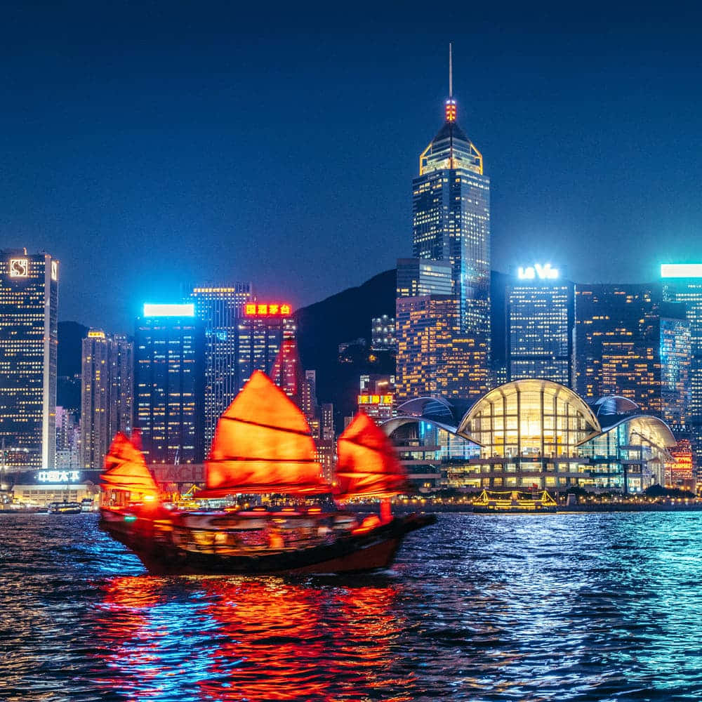 香港 桌布 壁紙 — 一艘傳統中式帆船，帶有發光的紅色船帆，在夜間滑過港口，反射著高聳摩天大樓的霓虹燈光。