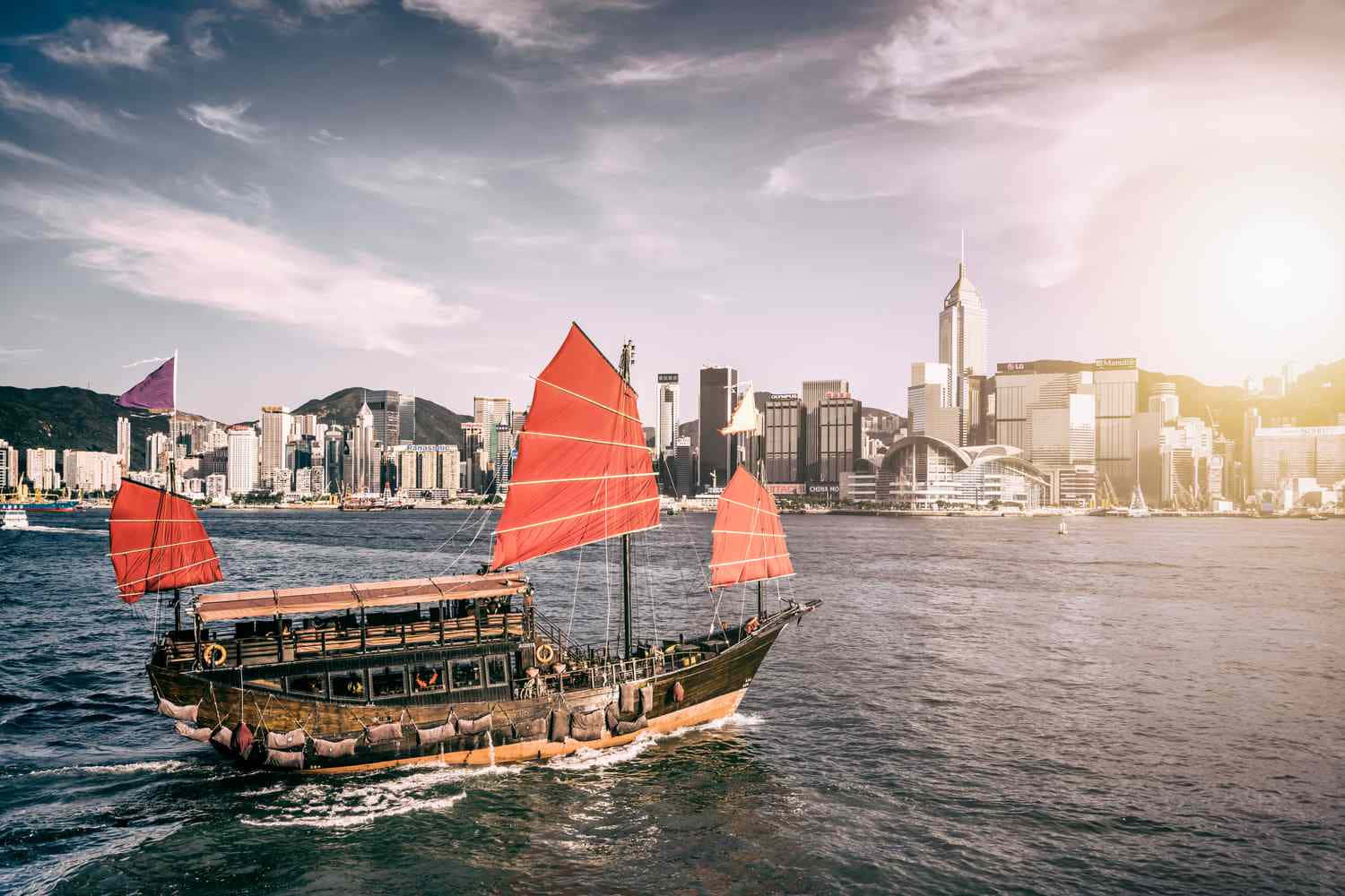 香港 桌布 壁紙 — 一艘傳統中式帆船，紅色船帆，在陽光照耀的水面上滑行，背景是香港高聳的天際線。