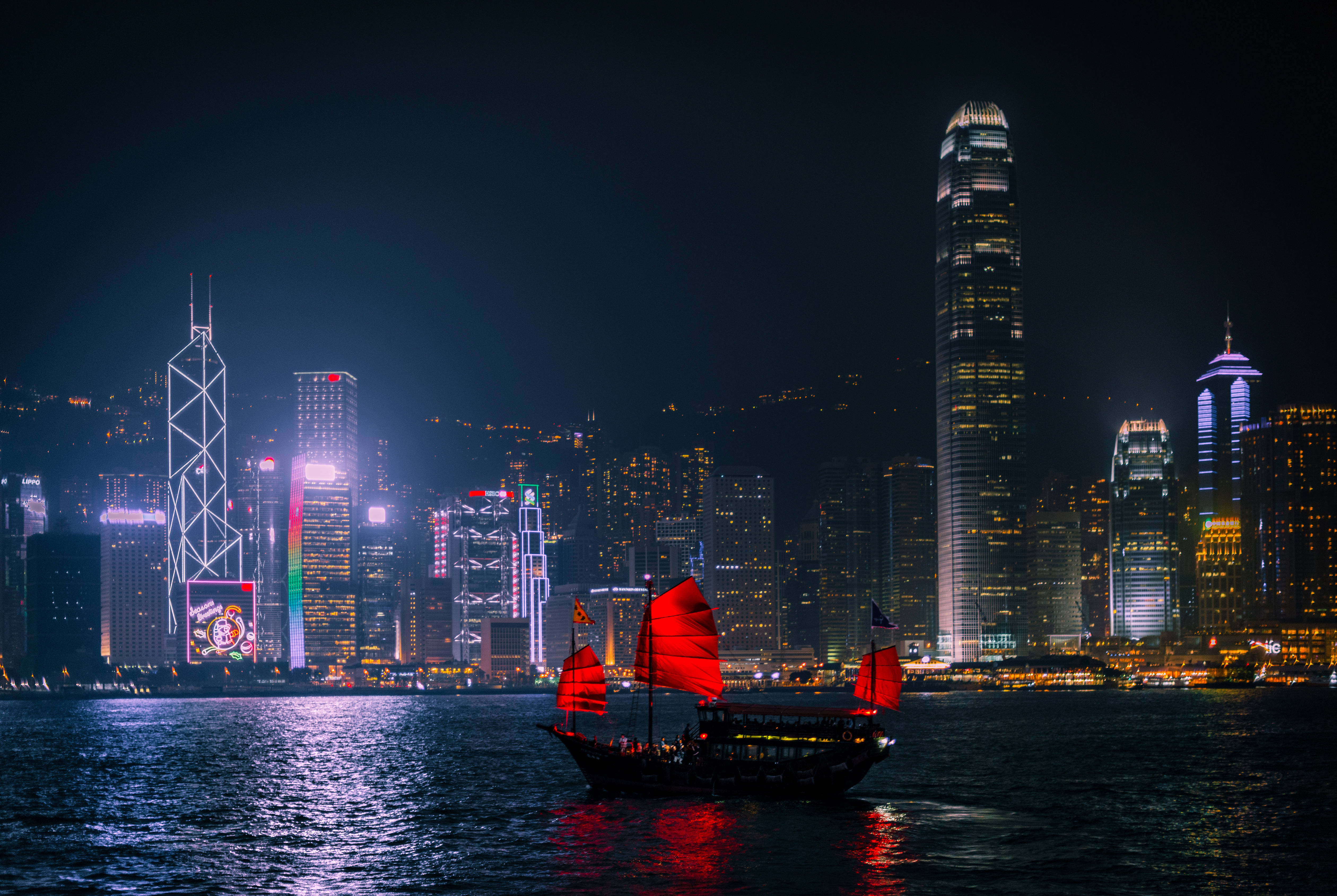 香港 桌布 壁紙 — 一艘傳統中式帆船帶著發光的紅帆，在夜間香港高聳天際線的耀眼霓虹燈下，滑過黑暗的港口。