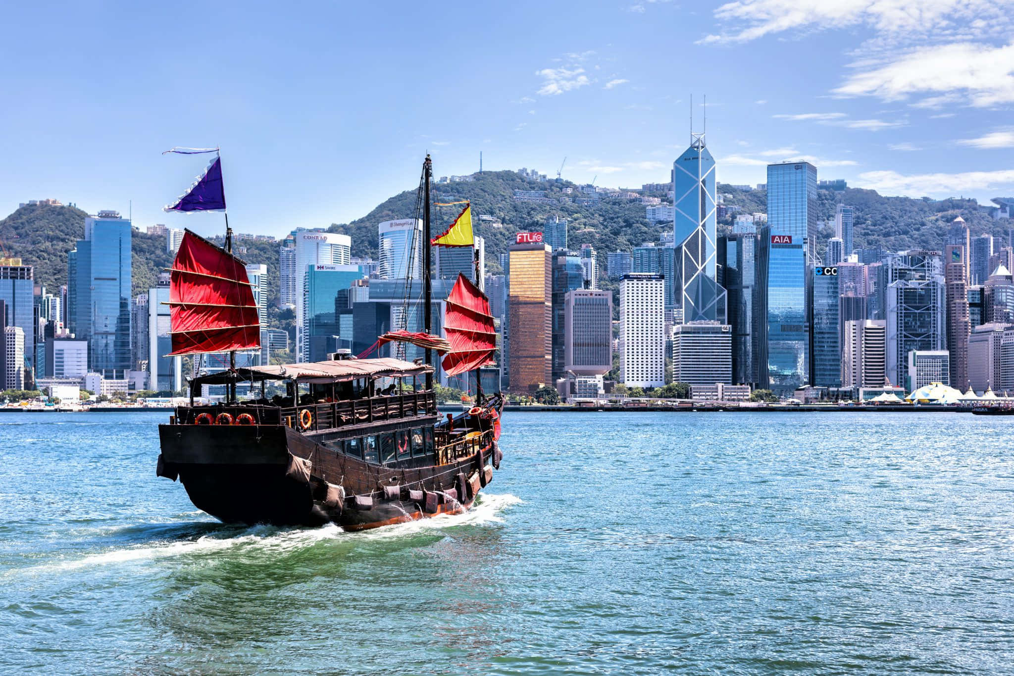 Hong Kong 桌布 壁紙 — 一艘傳統中式帆船張開紅帆，在碧綠色的海面上滑行，前方是香港高聳的玻璃摩天大樓，天空湛藍明亮。