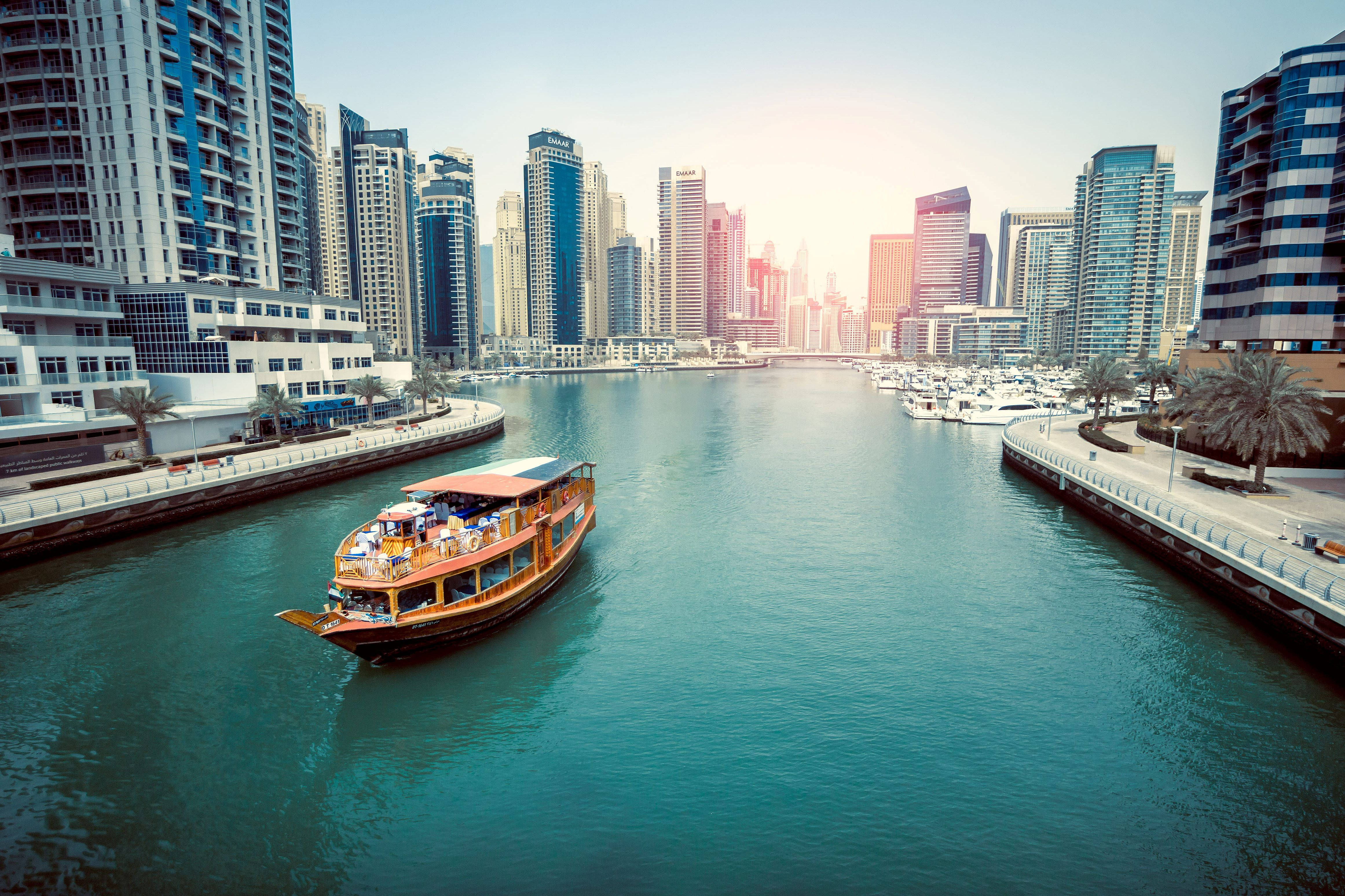 Arab 桌布 壁紙 — 一艘傳統木製單桅帆船遊艇在明亮的陽光下滑過一條碧綠色的運河，兩旁是高聳的現代摩天大樓。