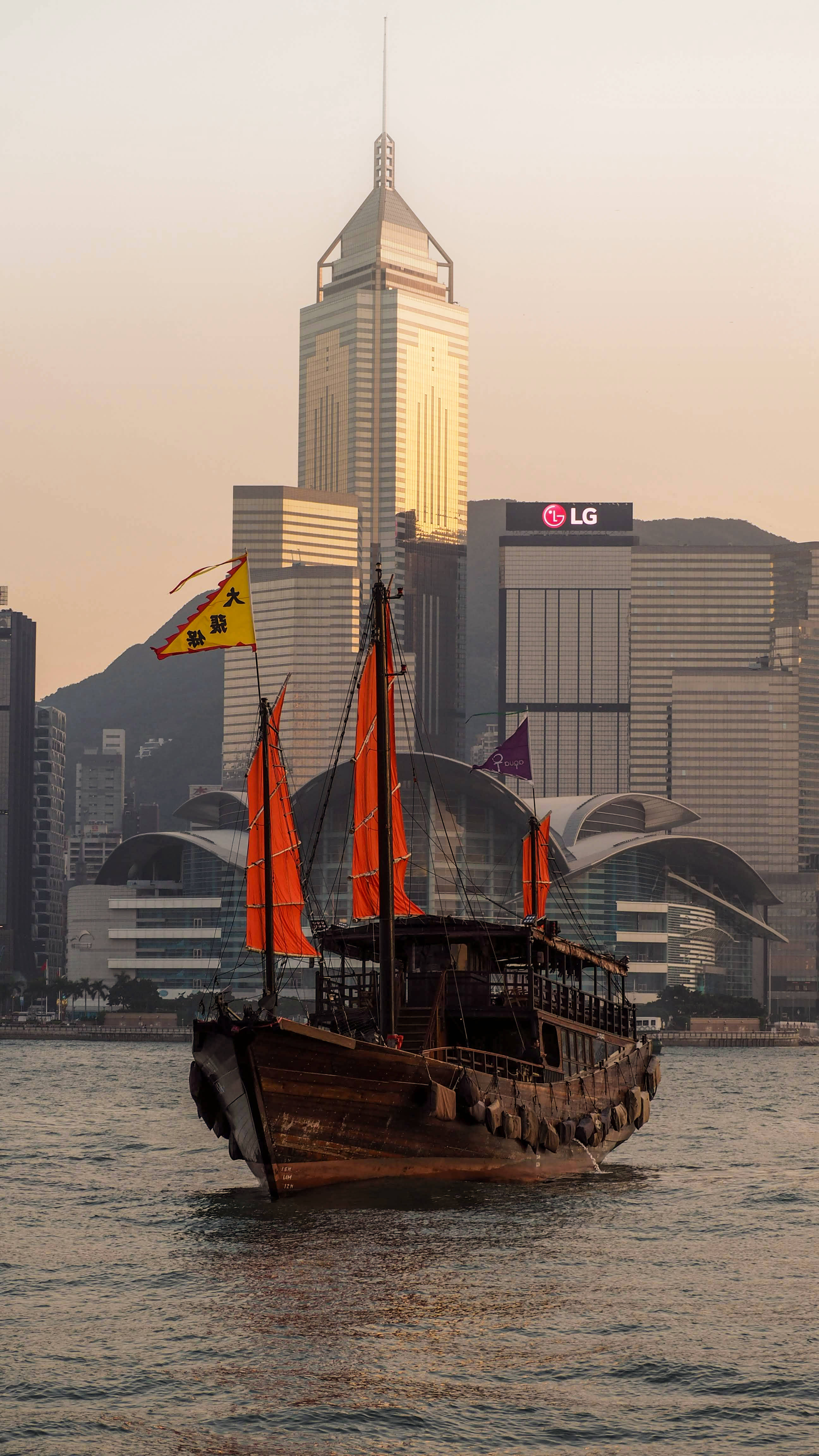 Hong Kong 桌布 壁紙 — 一艘傳統木製帆船，掛著橙色風帆，在黃金時段於香港高聳天際線前的平靜水面上滑行。