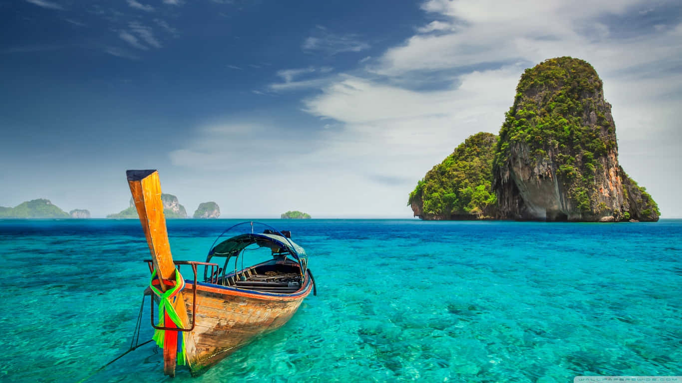 Thailand 桌布 壁紙 — 一艘傳統木製長尾船在清澈碧綠的海水中漂浮，靠近鬱鬱蔥蔥的綠色喀斯特島嶼，天空湛藍明亮。