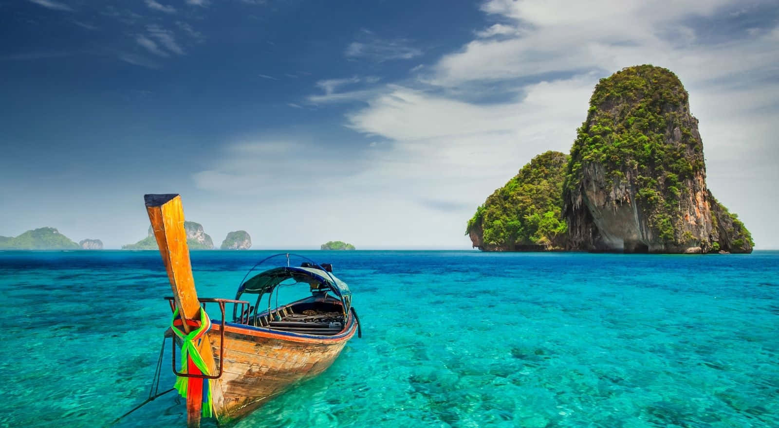 Travel Desktop 桌布 壁紙 — 一艘傳統木製長尾船漂浮在清澈碧綠的海水中，靠近一個鬱鬱蔥蔥的綠色喀斯特島嶼，天空湛藍明亮。