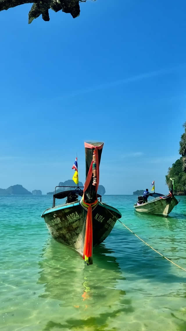 Cute Beach iPhone 桌布 壁紙 — 一艘傳統泰國長尾船在清澈碧綠的海水中休憩，背景是湛藍的天空和遠處的島嶼。