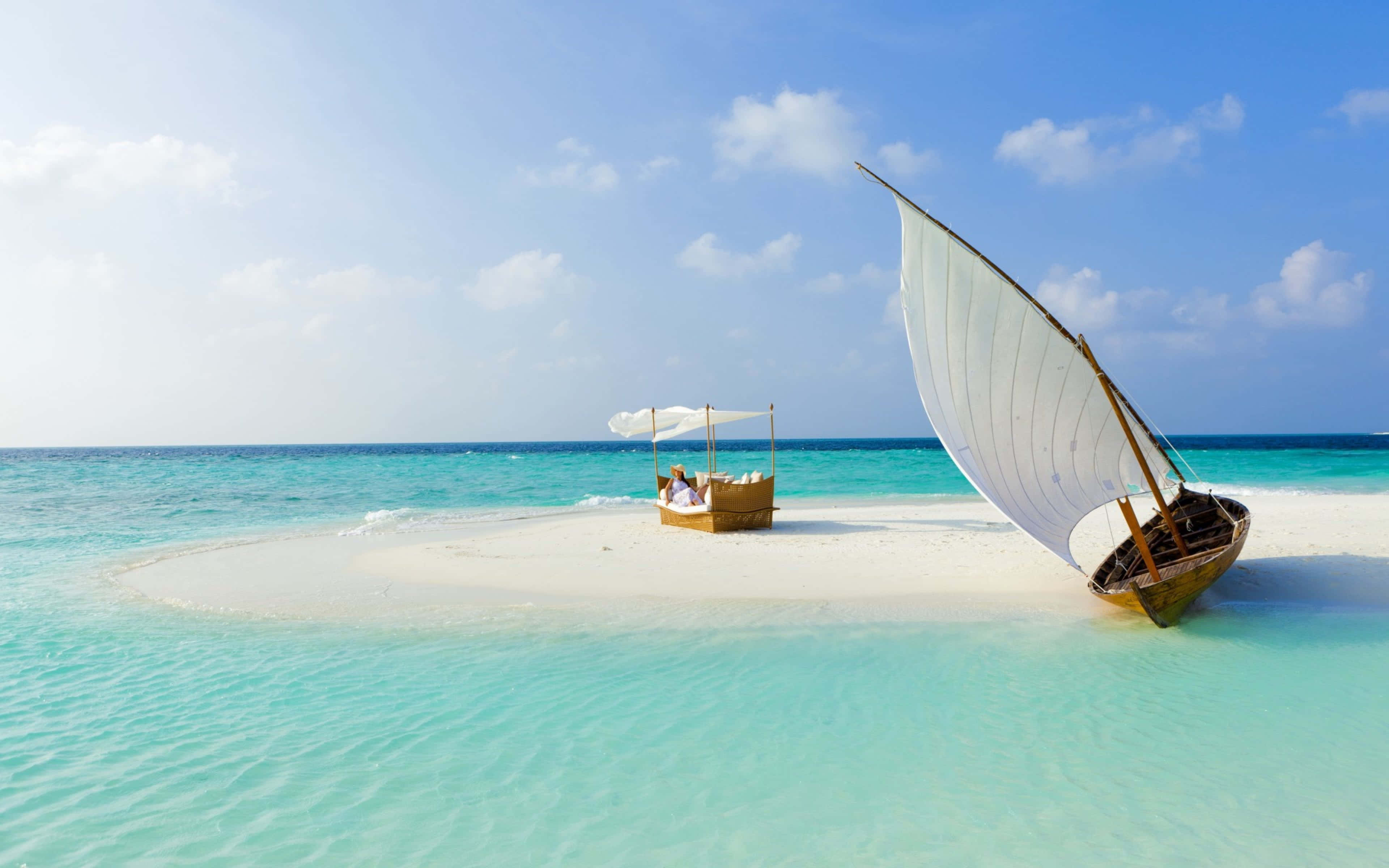 Beach Daybed 桌布 壁紙 — 一艘傳統的單桅帆船停泊在 pristine 白色沙洲上，四周環繞著清澈碧綠的海水，上方是明亮的藍天。