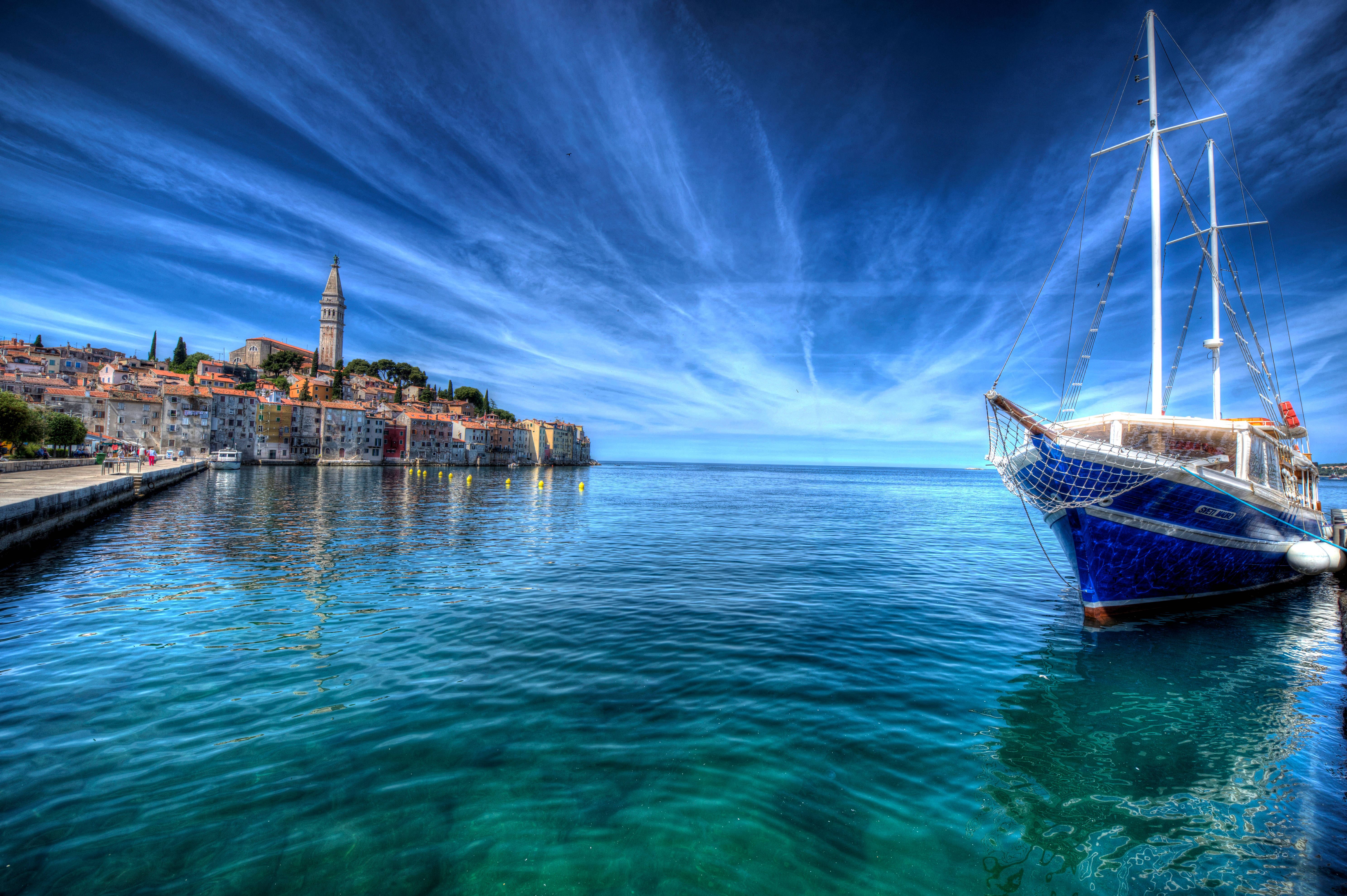 Croatia 桌布 壁紙 — 一艘充滿活力的藍色帆船停泊在清澈碧綠的海水中，旁邊是一個色彩繽紛的沿海小鎮，天空壯麗。